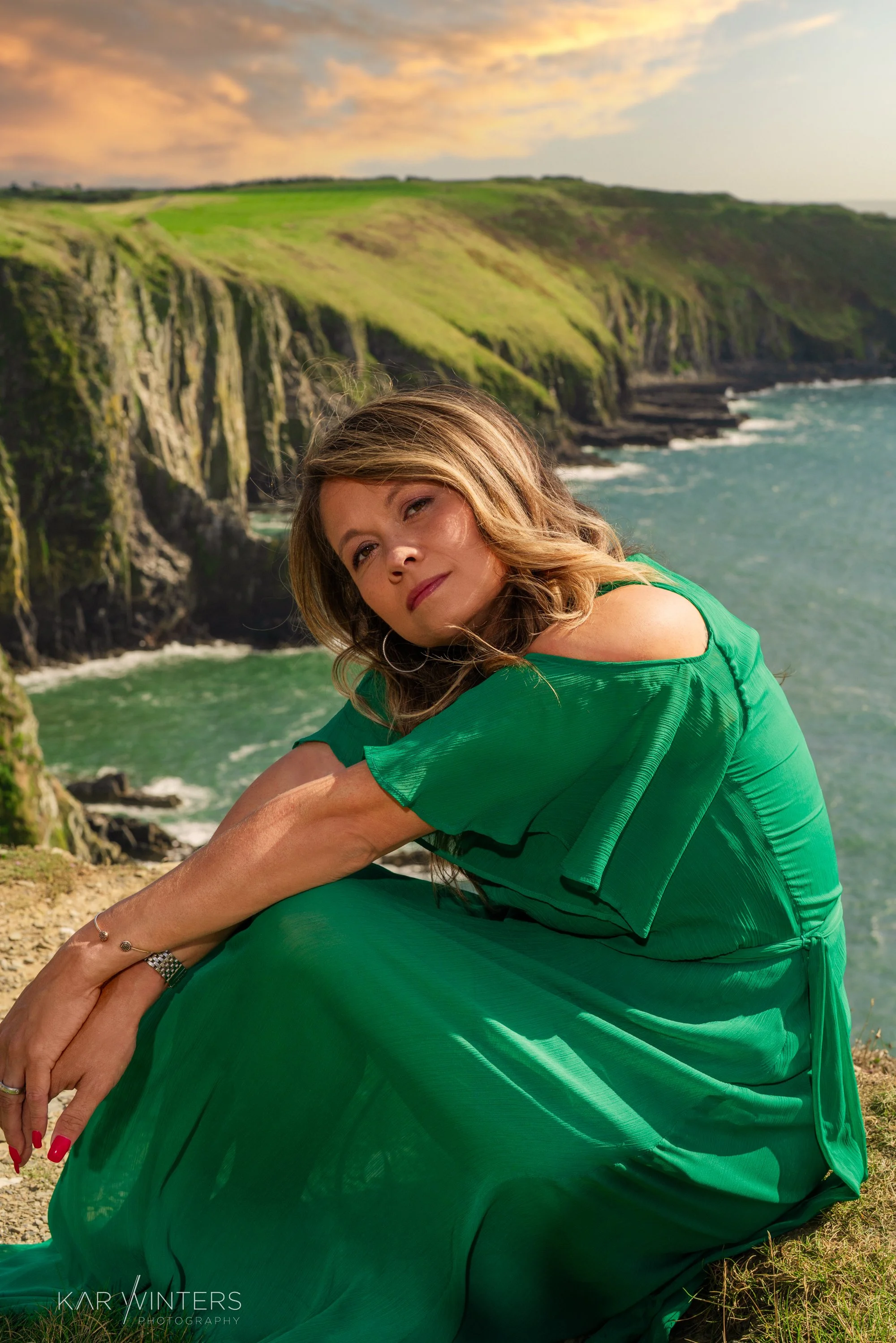 A woman in a green dress sitting on a grassy cliff, with the ocean and green cliffs in the background during sunset.