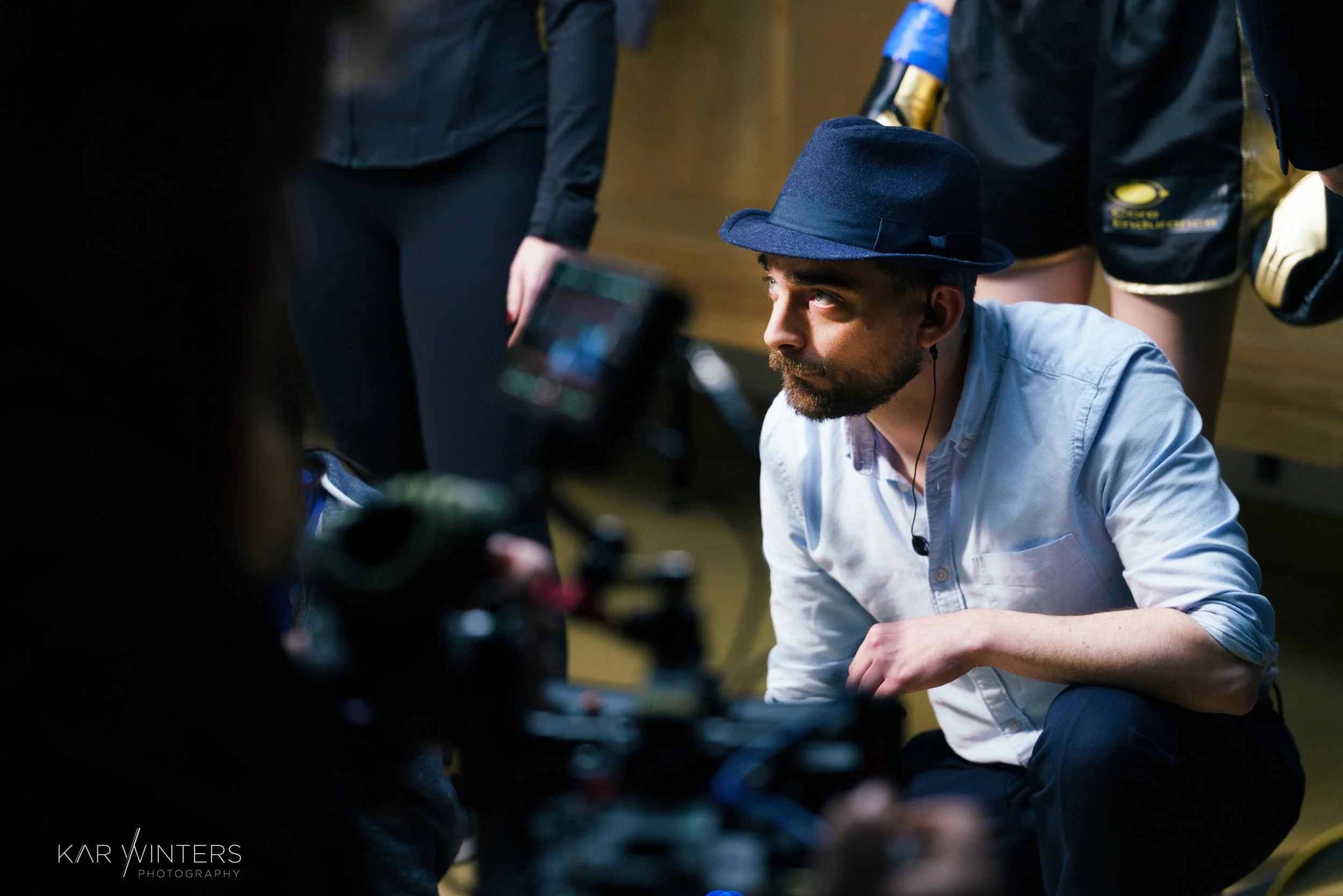 A man with a beard and wearing a blue fedora hat and a light blue shirt sitting on the floor surrounded by people, looking up and to his left during a filming or photography session.