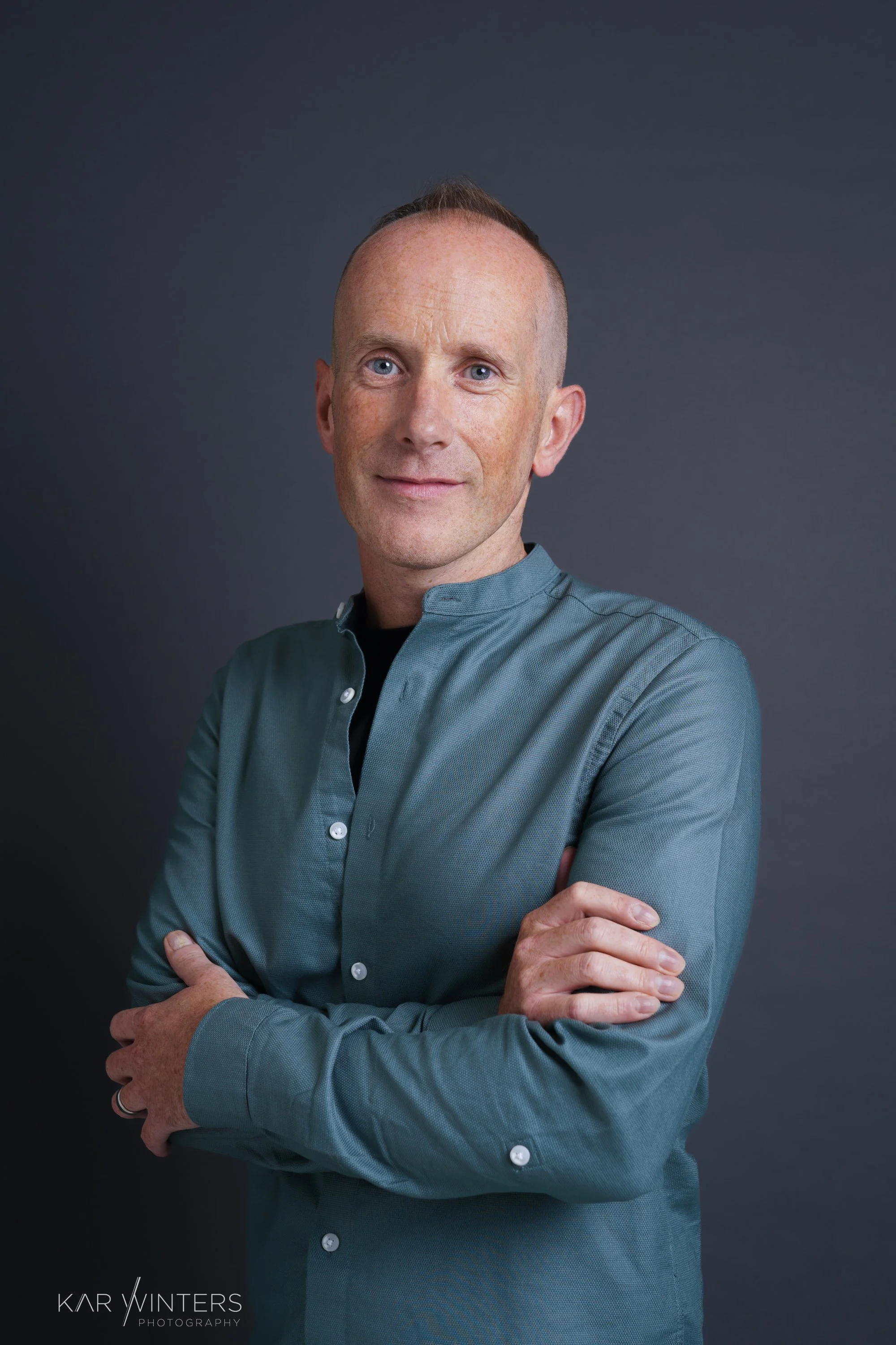 Portrait of a man with light skin, blue eyes, and receding hair, wearing a teal button-up shirt with arms crossed, standing against a dark gray background.