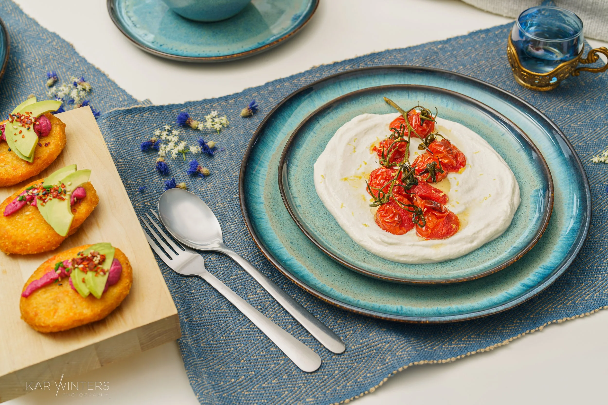 A table setting with a blue and turquoise platter of whipped feta with cherry tomatoes, a small glass of water, and three breaded appetizers topped with avocado slices and colorful sprinkles, on a wooden serving board. The table has a blue woven plac