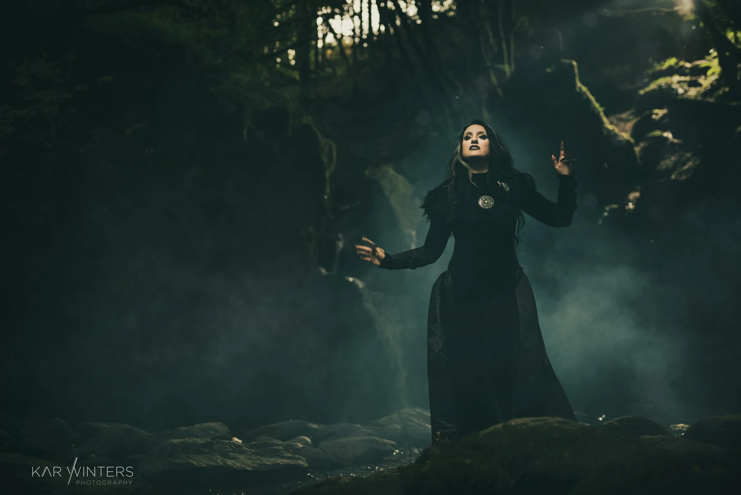 A woman dressed in gothic attire standing in a foggy forest, with her arms raised and dark makeup.