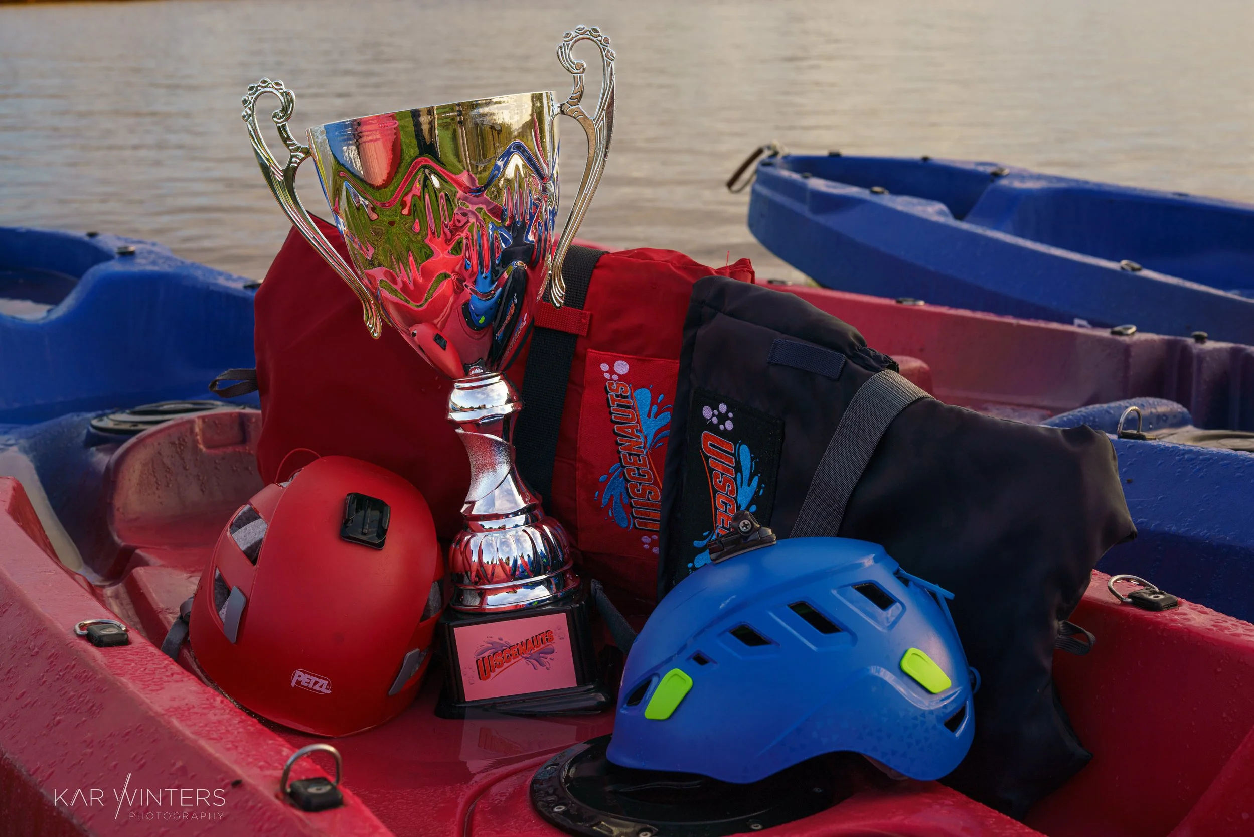 A trophy, two helmets, and gear bags placed in a red kayak on water.
