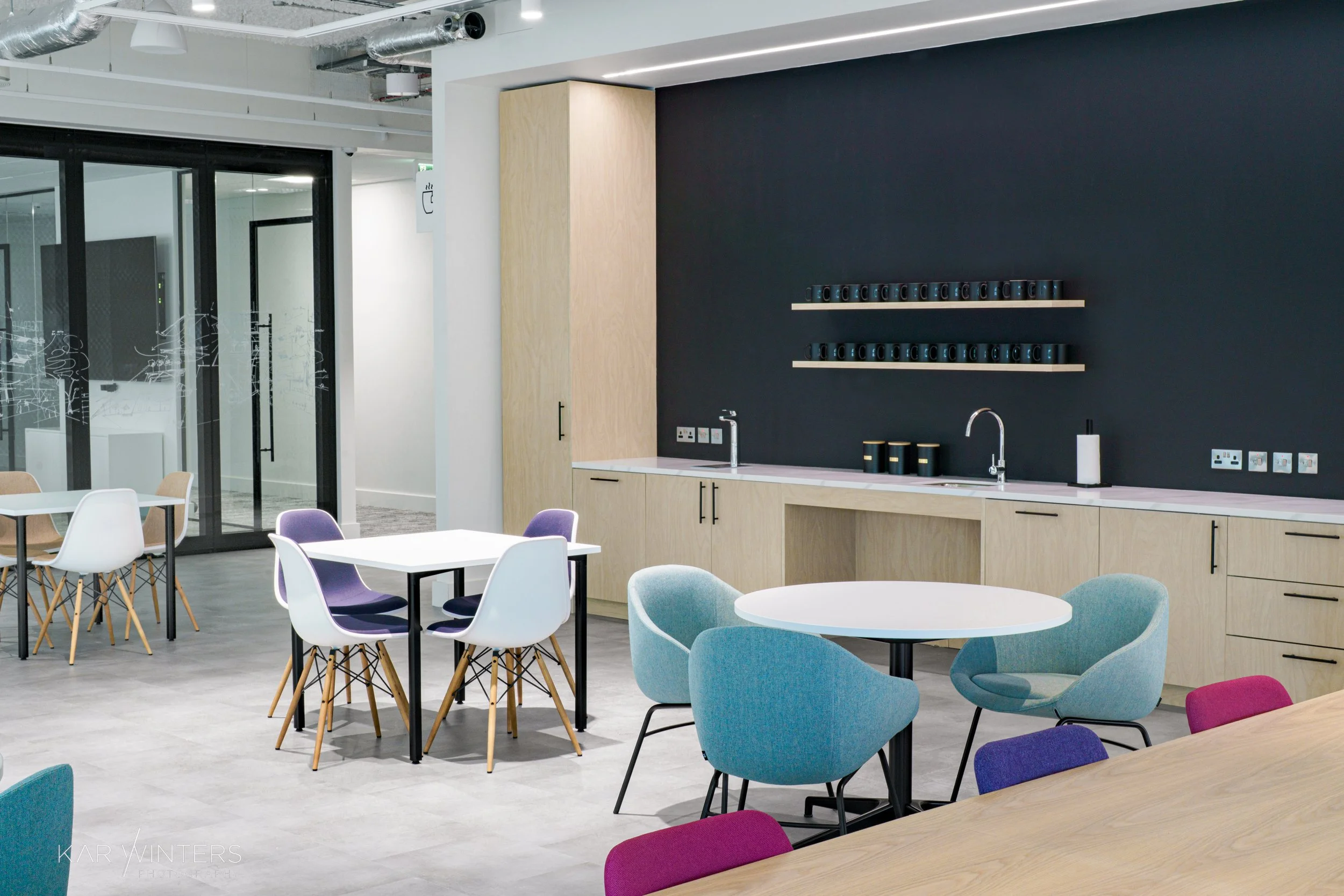 Modern kitchen break room with light wood cabinetry, black wall, floating shelves holding cups, two sinks, and colorful chairs around round and square tables.