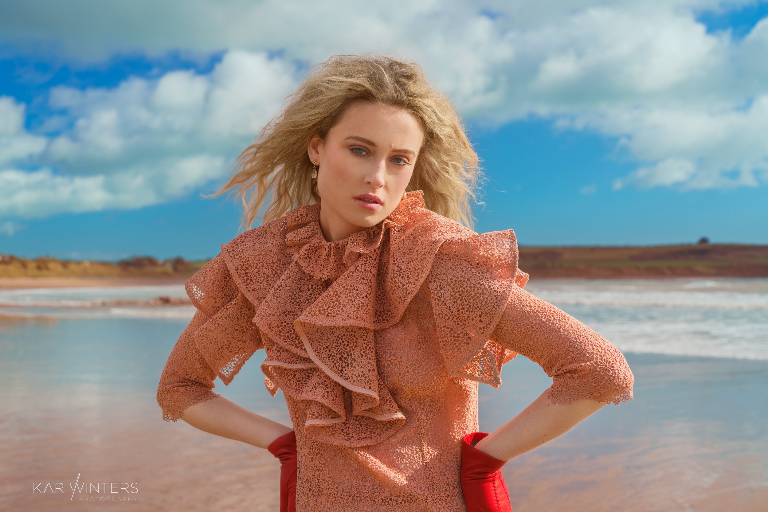 A woman with blonde hair standing on a beach, wearing a peach-colored lace dress with ruffles and red gloves, under a partly cloudy sky.