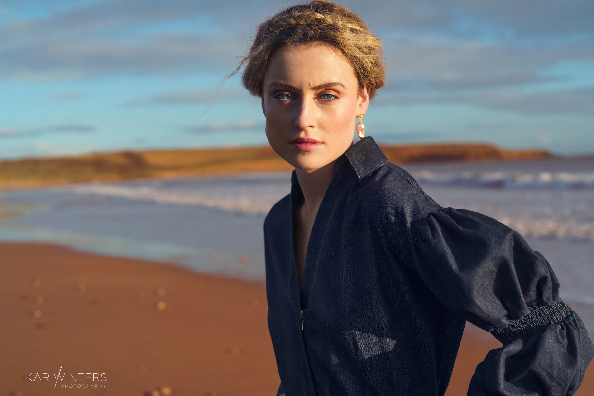 A woman with blue eyes and blonde hair styled in an updo, wearing a black jacket and gold earrings, standing on a beach with waves and sky in the background.