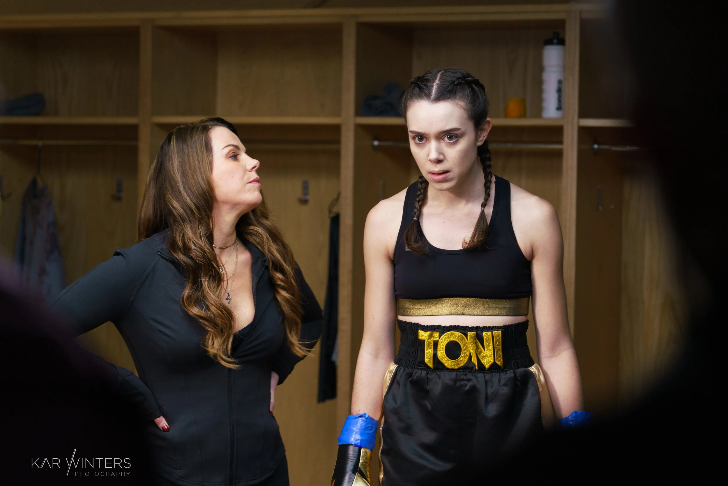 A young female boxer with braided hair, wearing a black boxing outfit with 'TONI' on the waistband, stands in a locker room with a worried expression. An older woman with long brown hair stands nearby, looking at her with concern.