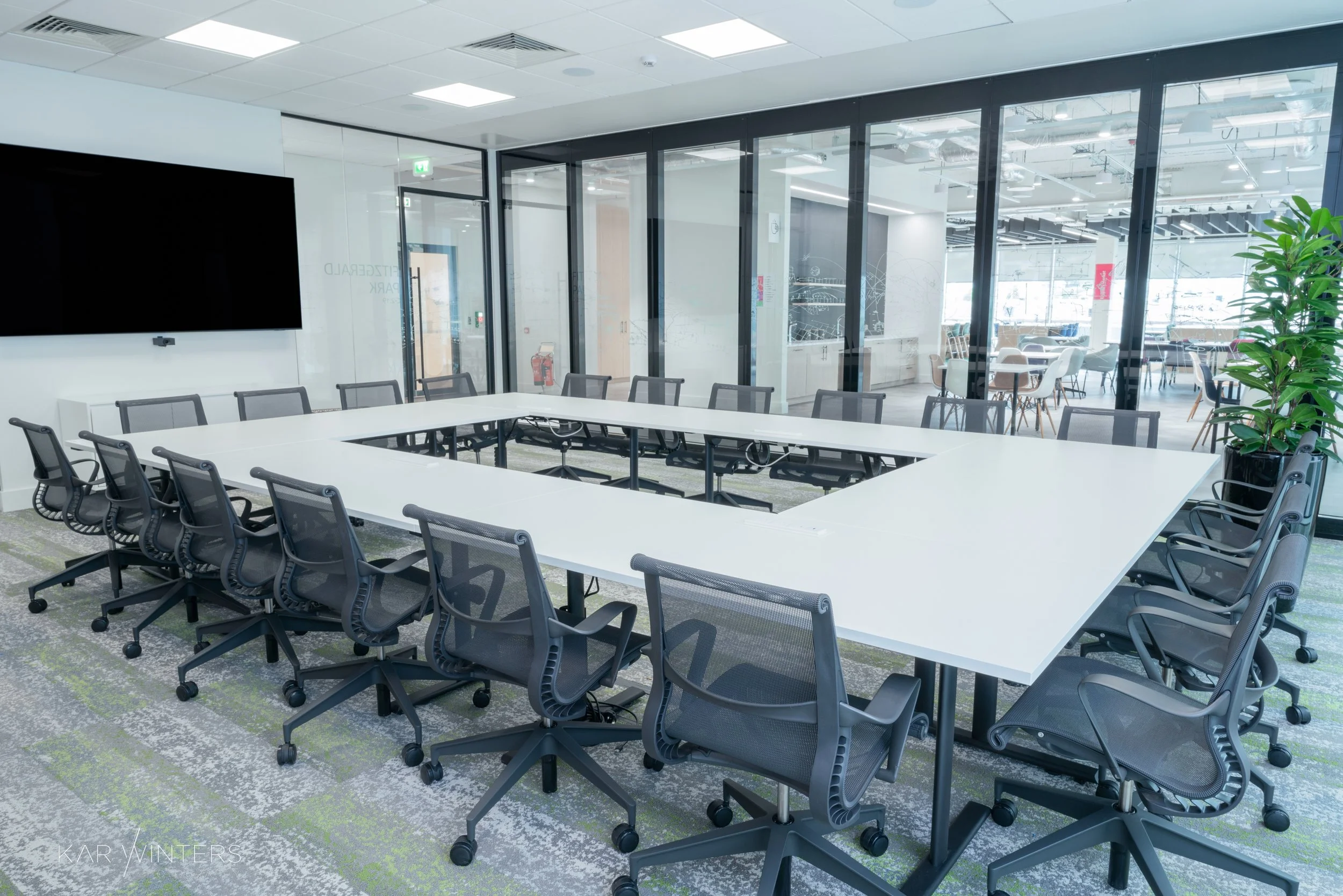 Empty modern conference room with U-shaped white table, black mesh office chairs, large black television, and glass walls with view into a lounge area with additional seating and natural light.