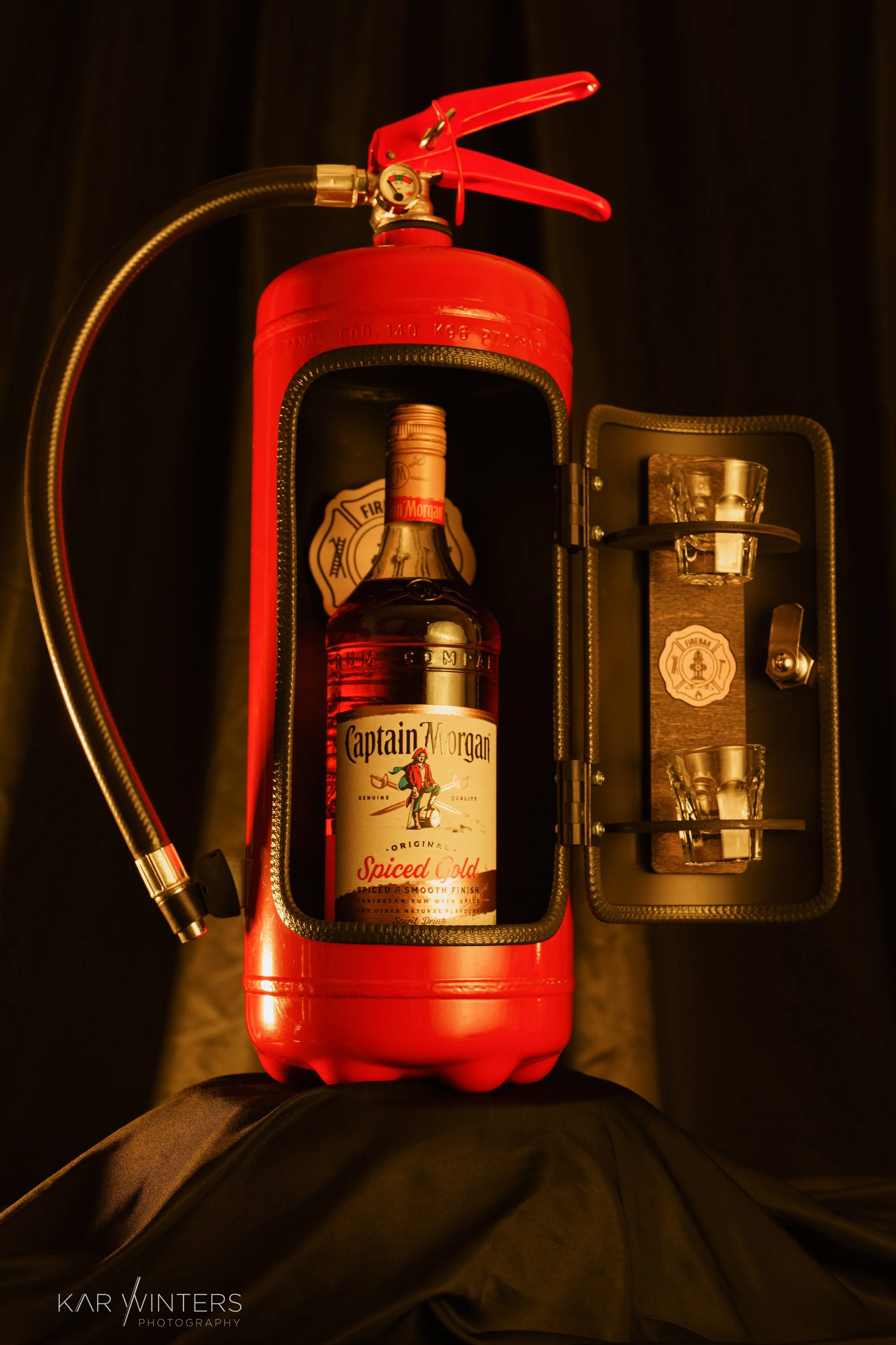 An antique fire alarm bell converted into a bar display, with a bottle of Captain Morgan Spiced Gold rum inside and shot glasses on a wall mount within the door.