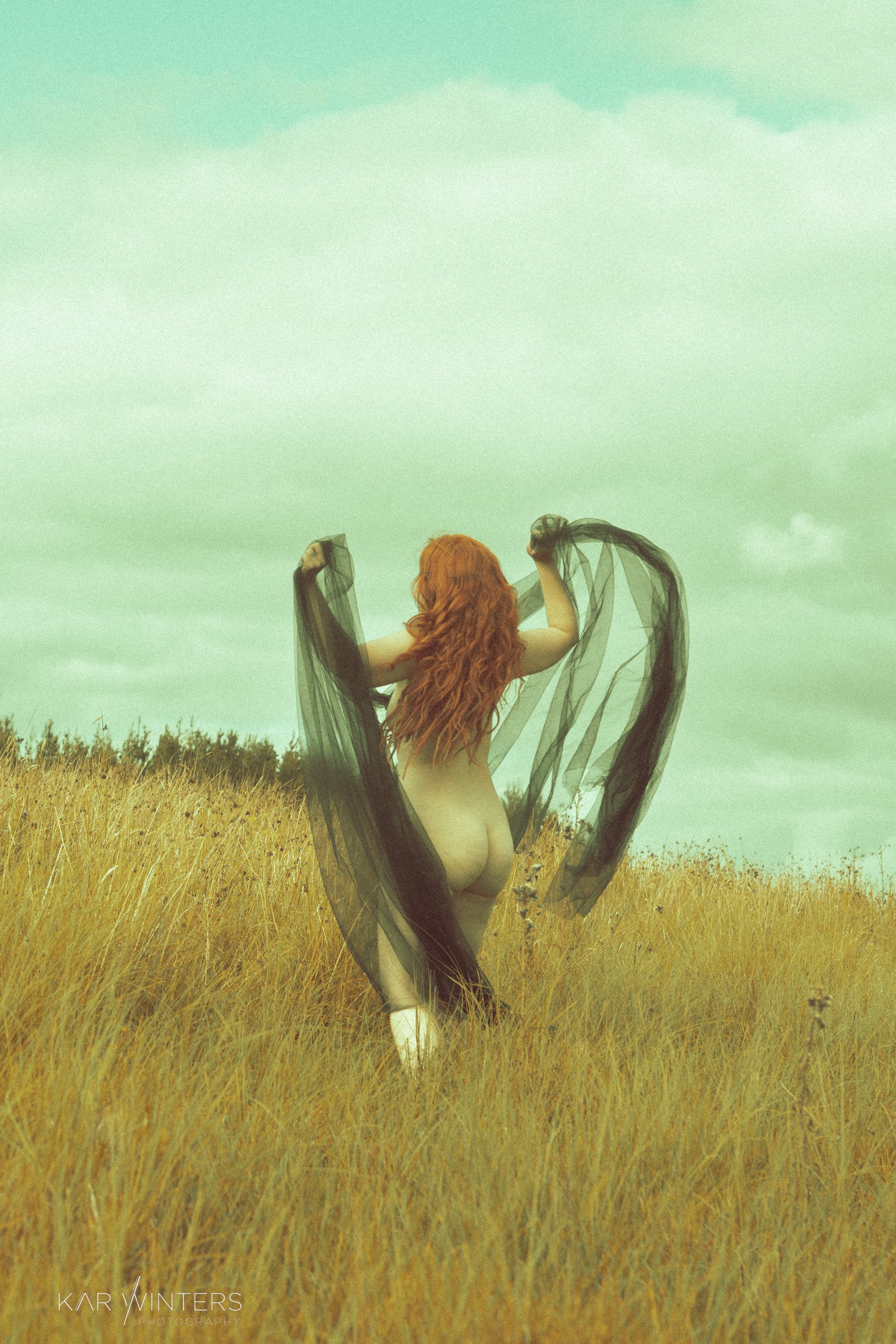 A woman with long red hair standing in a golden field holding black sheer fabric, facing away from the camera.