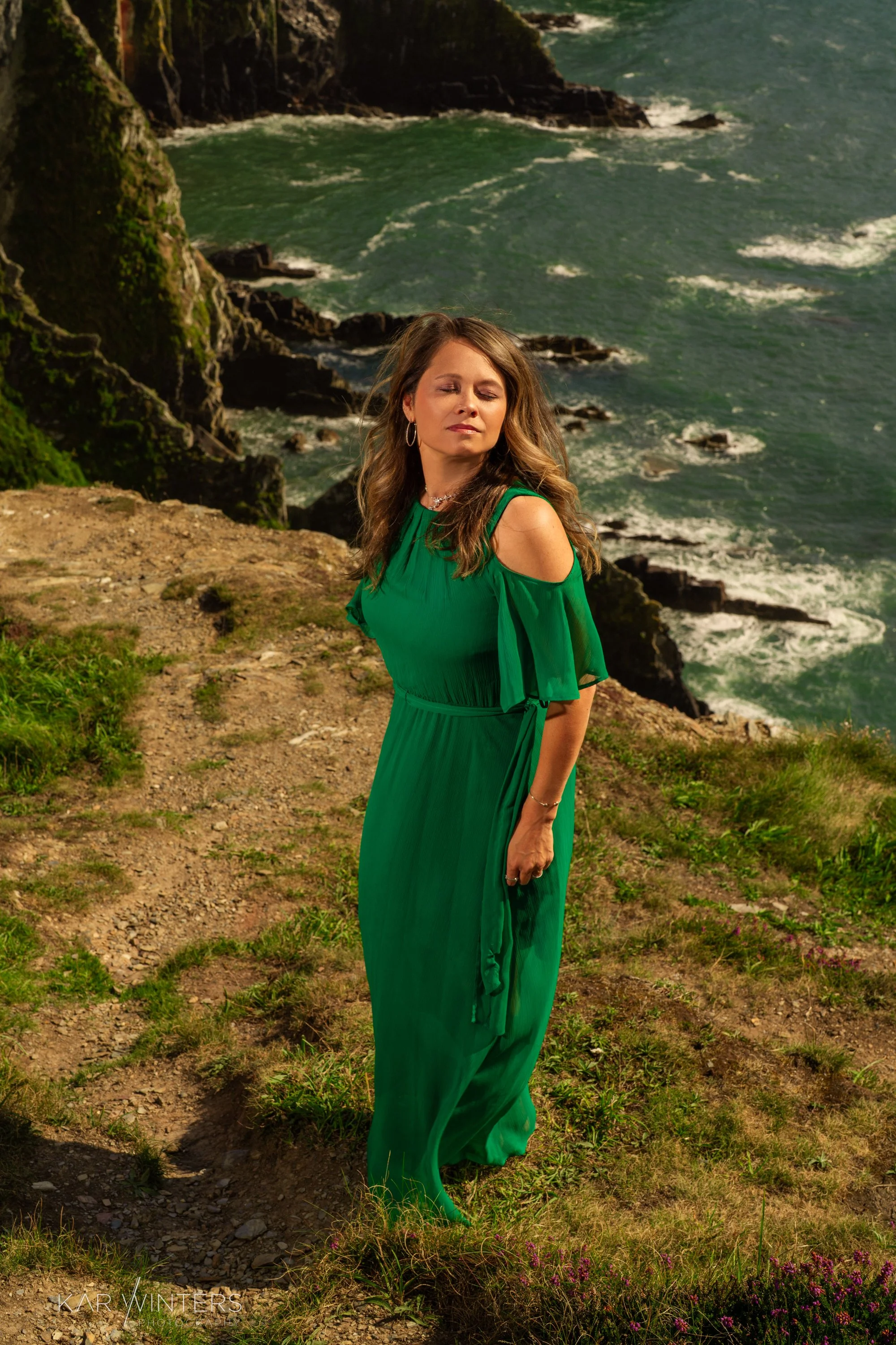 Woman in a green dress standing on a cliffside overlooking the ocean.
