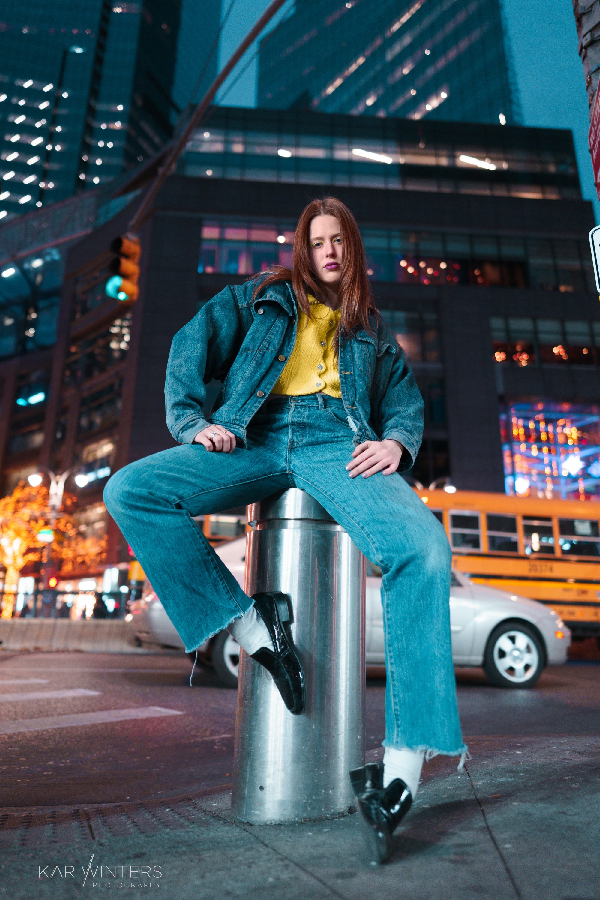 A young woman with red hair sitting on a street bollard in an urban area at night, with tall buildings lit up in the background.
