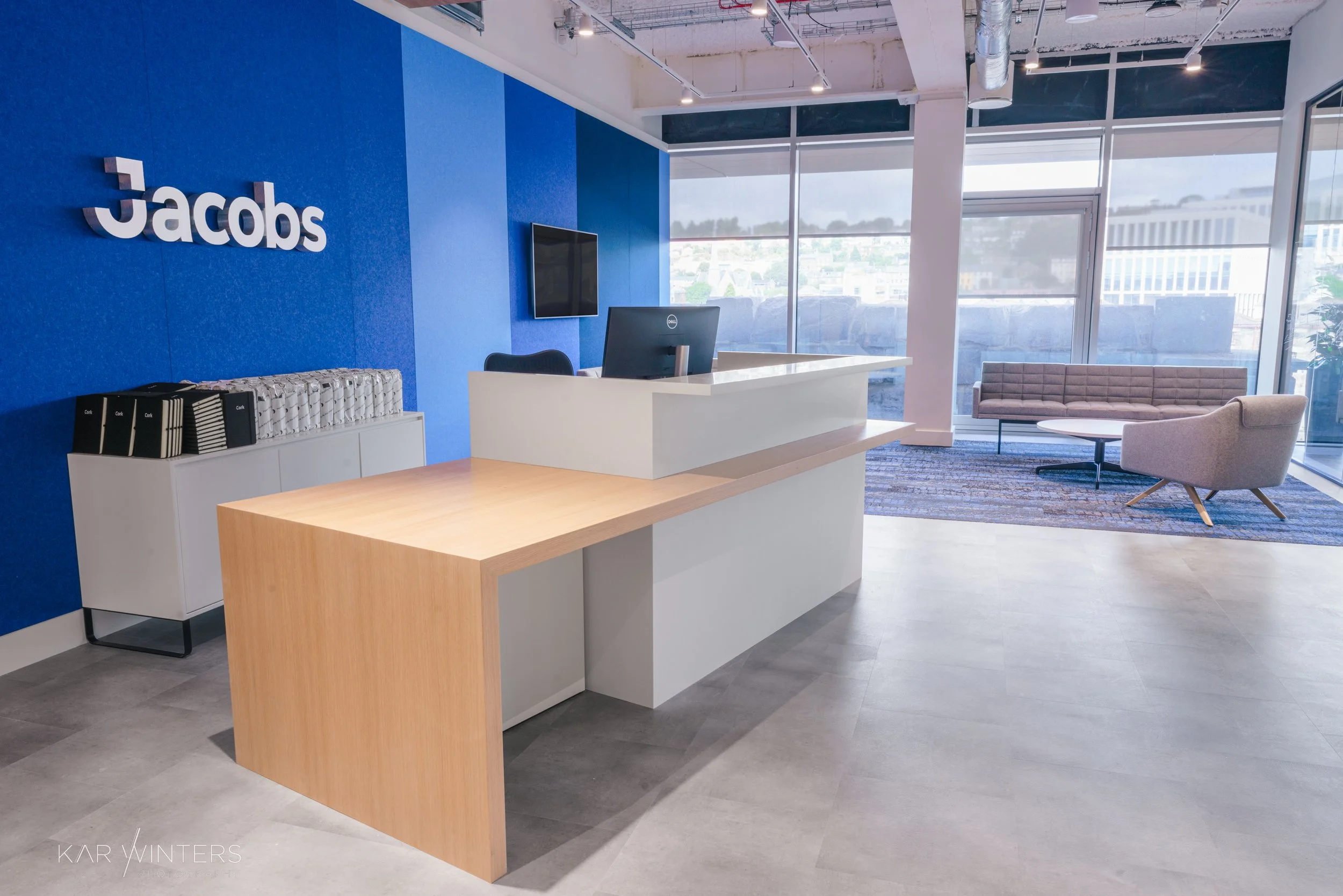 Modern office reception area with a white desk, a computer, a blue accent wall with the 'Jacobs' logo, and a lounge with chairs and a coffee table near large windows showing a city view.