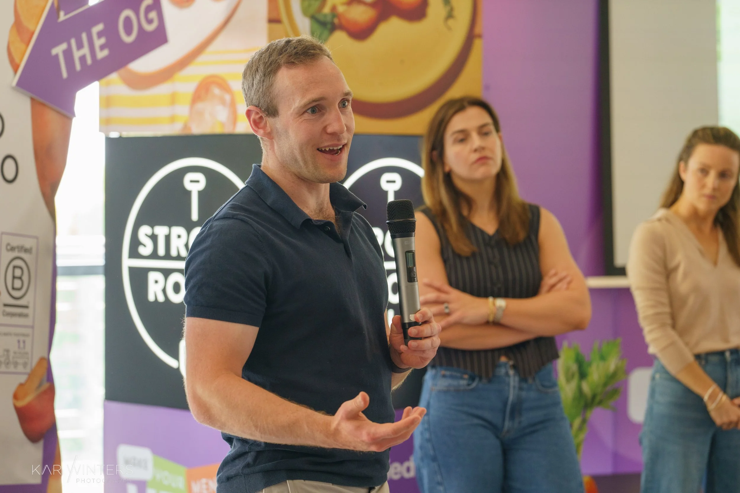 A man in a navy blue polo shirt speaking into a microphone during a presentation or event, with two women standing behind him, one crossing her arms and the other holding her hands in front of her, in a colorful indoor setting with banners and signs.
