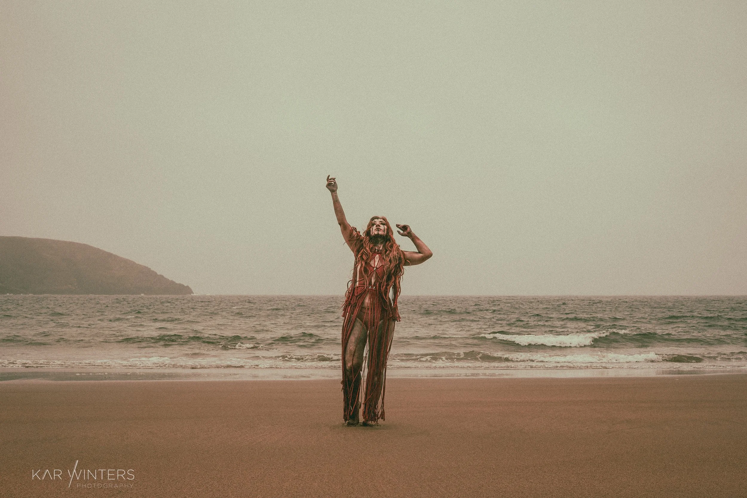 Woman with long hair in bohemian attire dancing on the beach near the ocean with an island in the background.