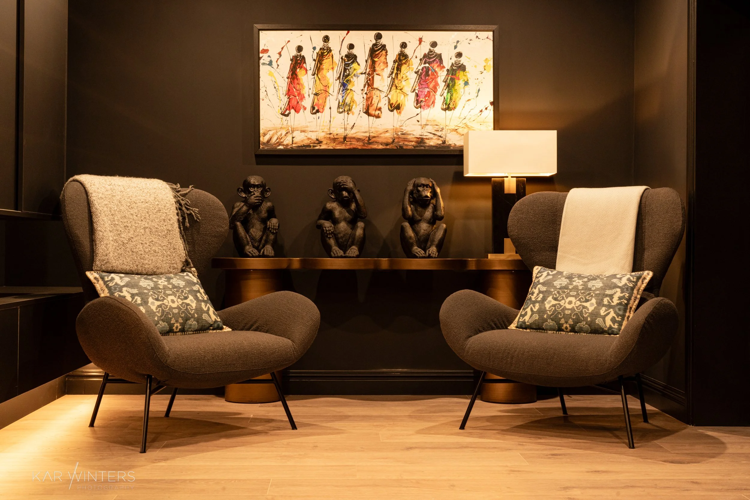 A cozy sitting area with two dark gray armchairs with patterned pillows, a wooden table in the background with three black monkey sculptures, a table lamp, and a colorful abstract painting on the dark wall.
