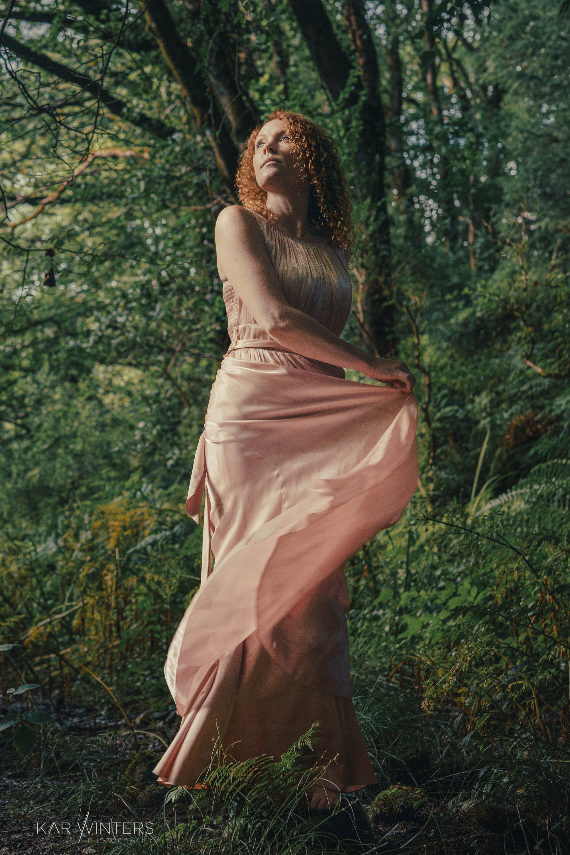 A woman with curly red hair in a flowing pink dress standing in a forest, looking upward.