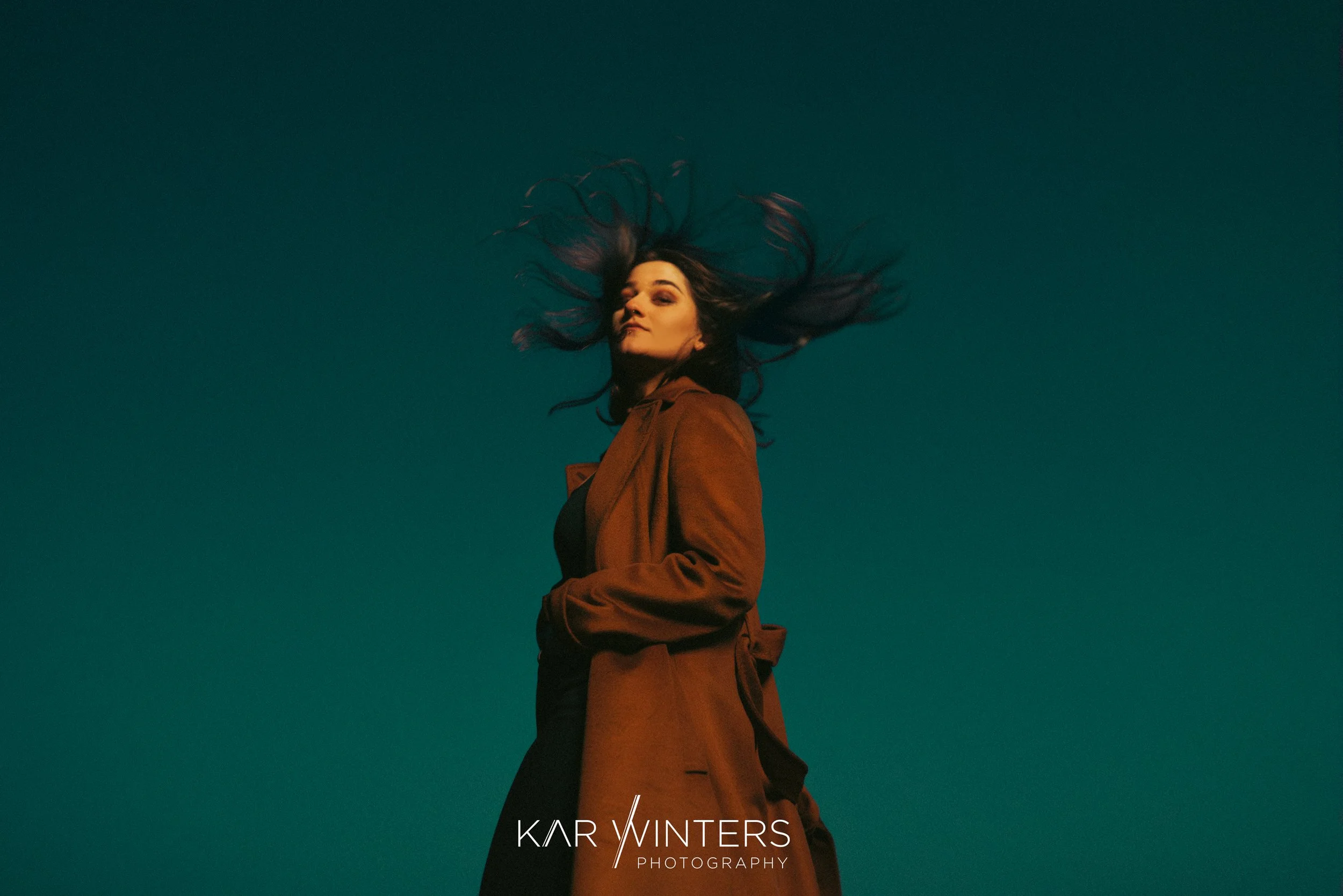 A woman with dark hair in motion against a dark teal sky, wearing a brown coat and black outfit, looking confident with her hair flowing outward.