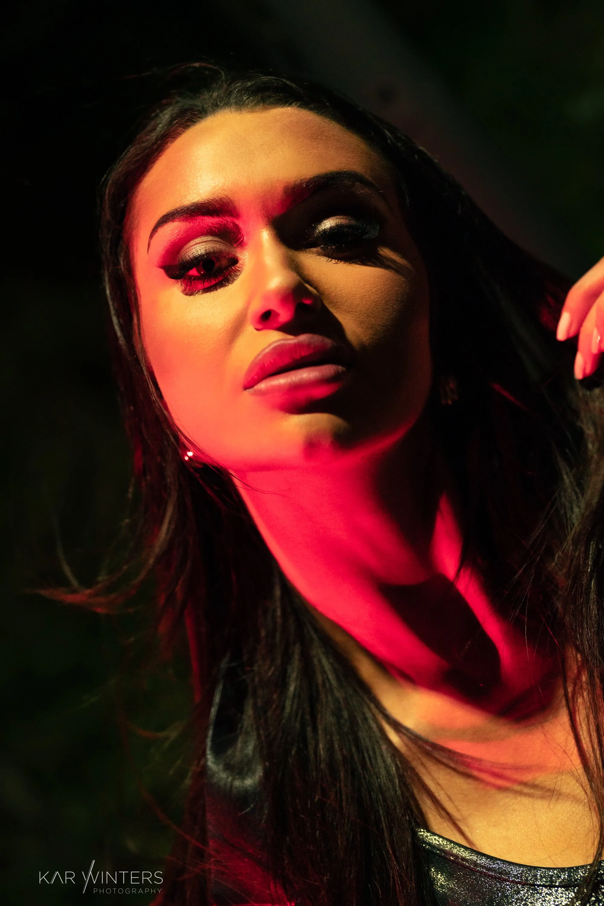 A woman with dark hair and makeup, illuminated by red and green lighting, looking directly at the camera.