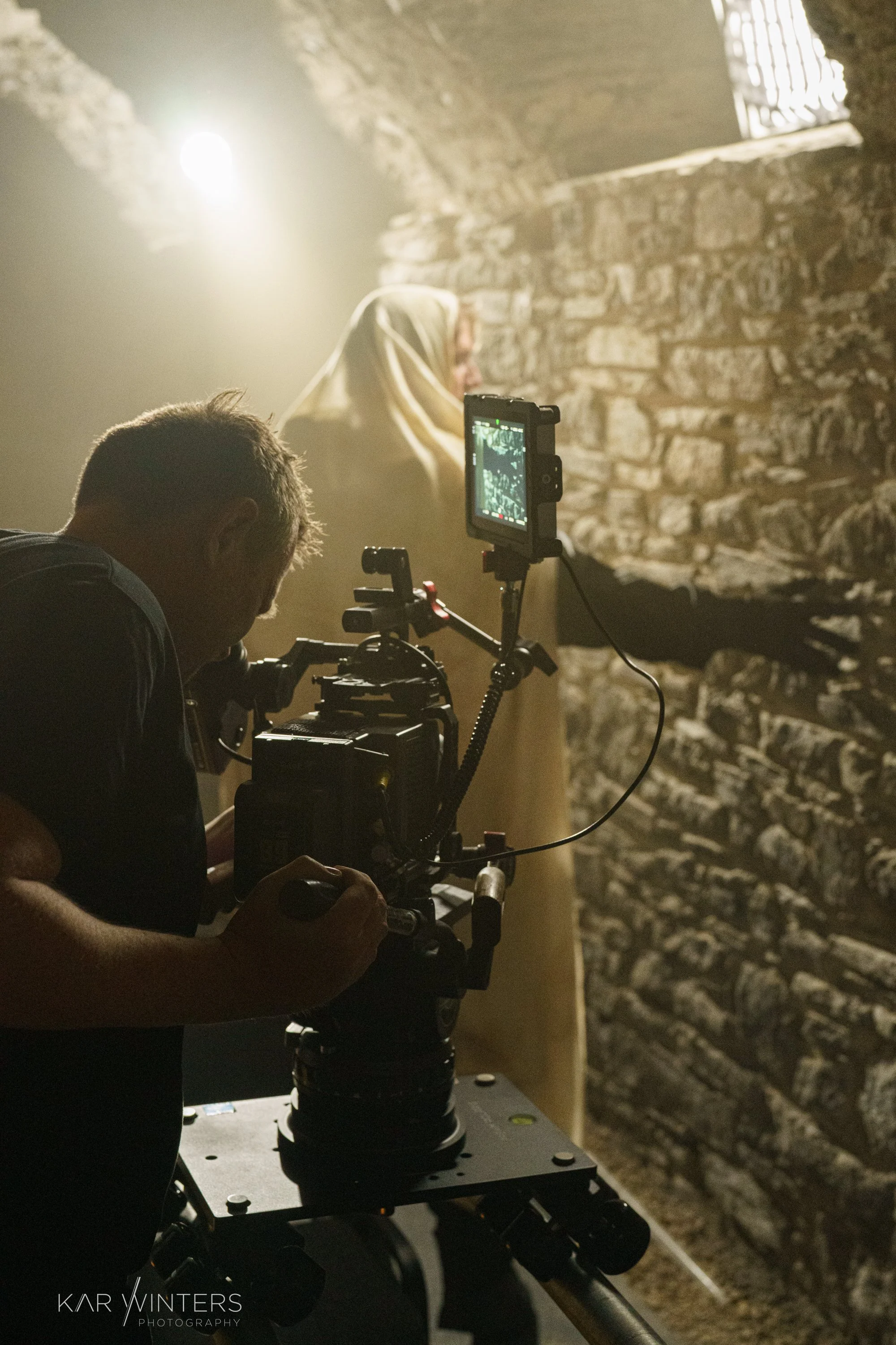 A film crew operating a camera in a dimly lit stone-walled corridor, filming a woman in traditional clothing with a beige headscarf.