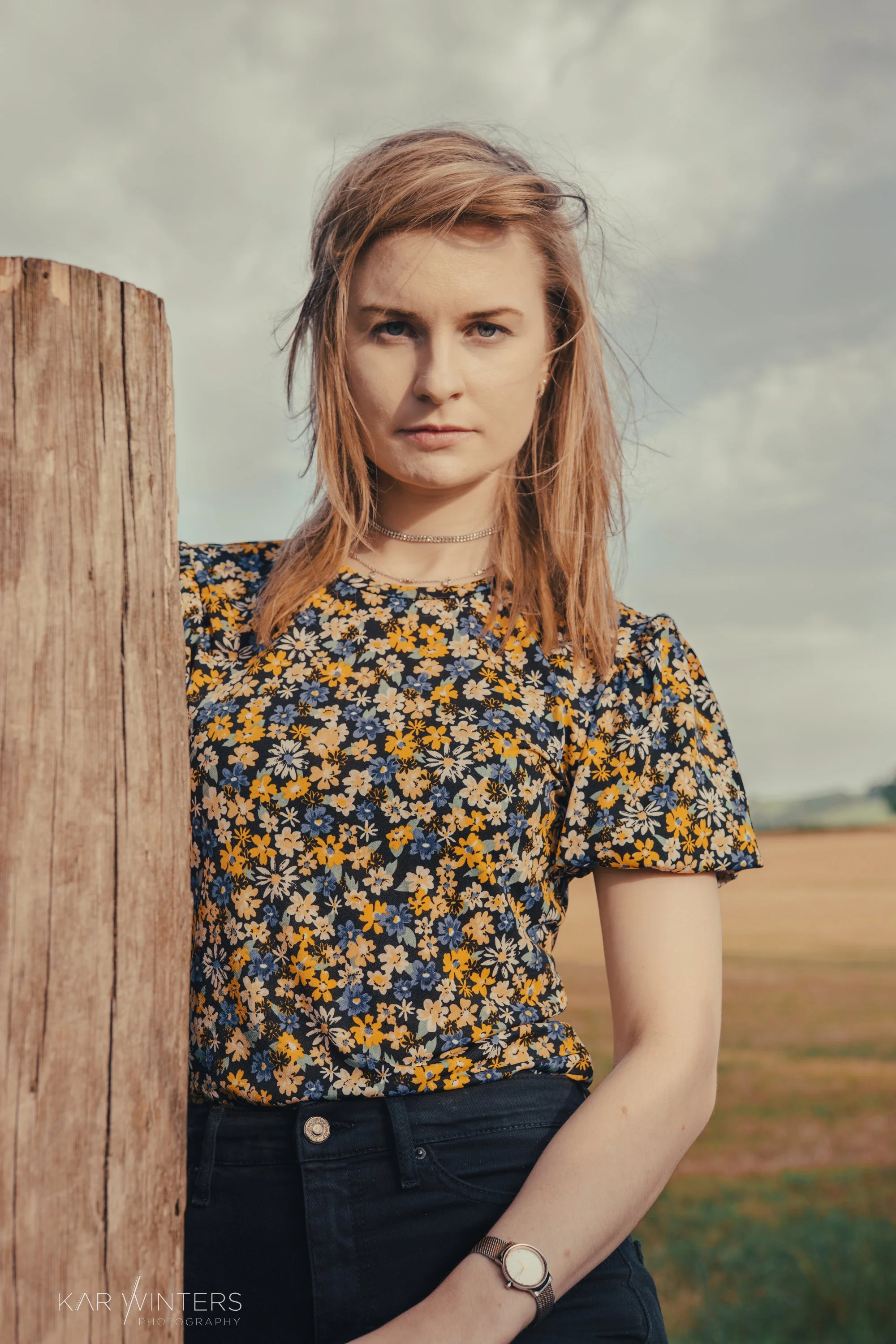 A young woman with light brown hair and blue eyes standing outdoors beside a wooden post, wearing a floral shirt and black jeans, with a cloudy sky in the background.