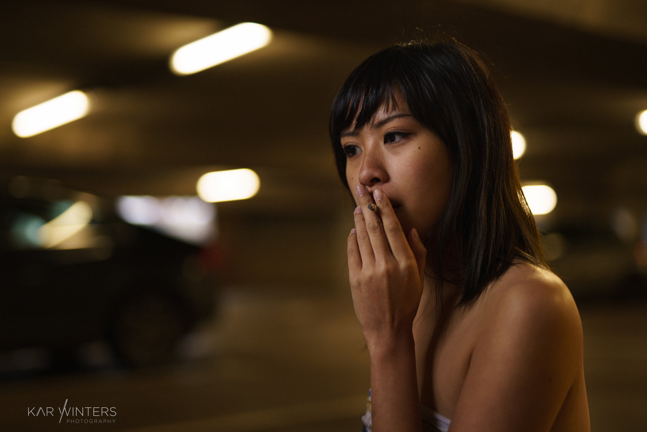 A young woman with short dark hair, topless, standing in an indoor parking garage with blurred lights in the background, touching her lips with her hand.
