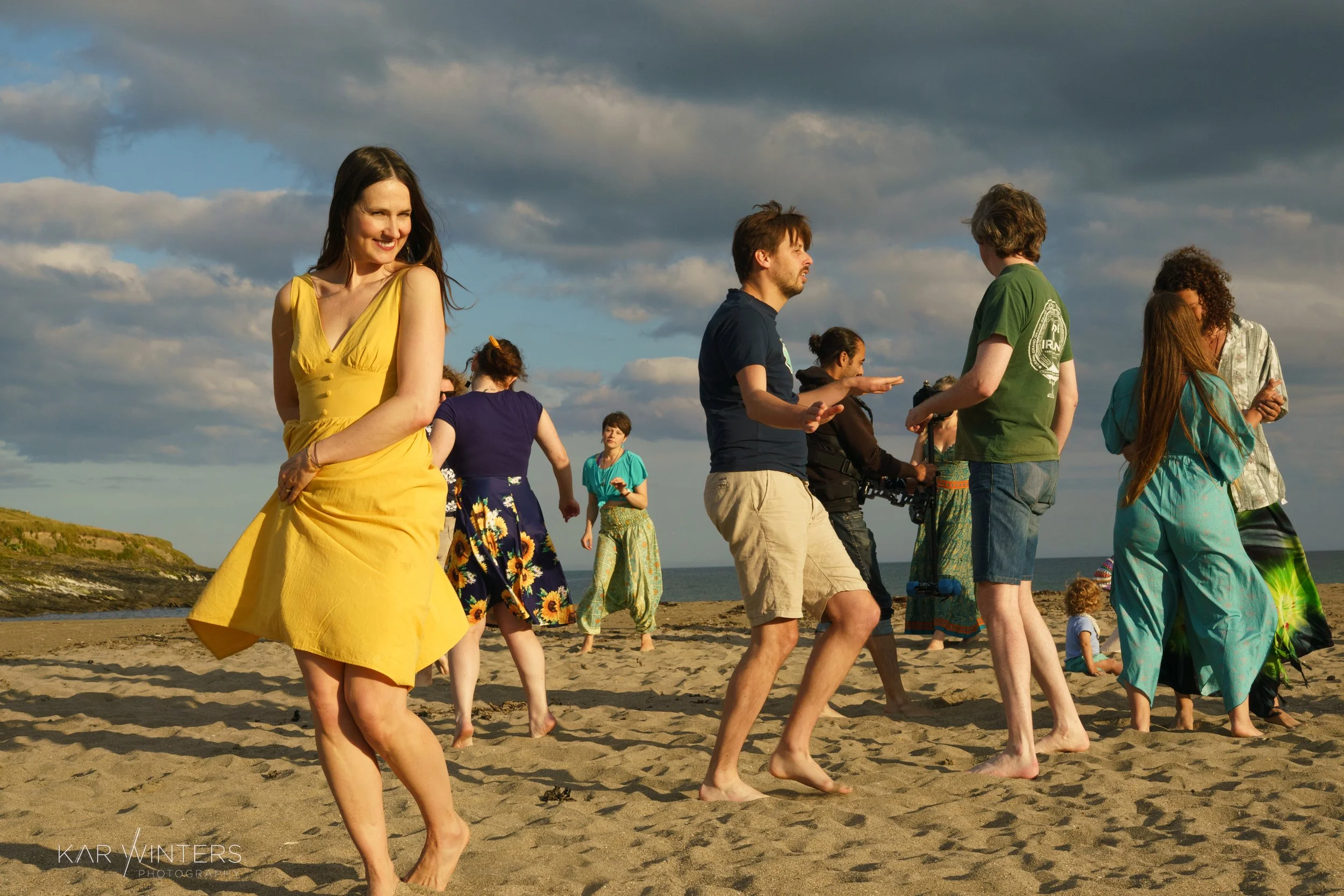 People dancing and socializing on a beach with a cloudy sky in the background.