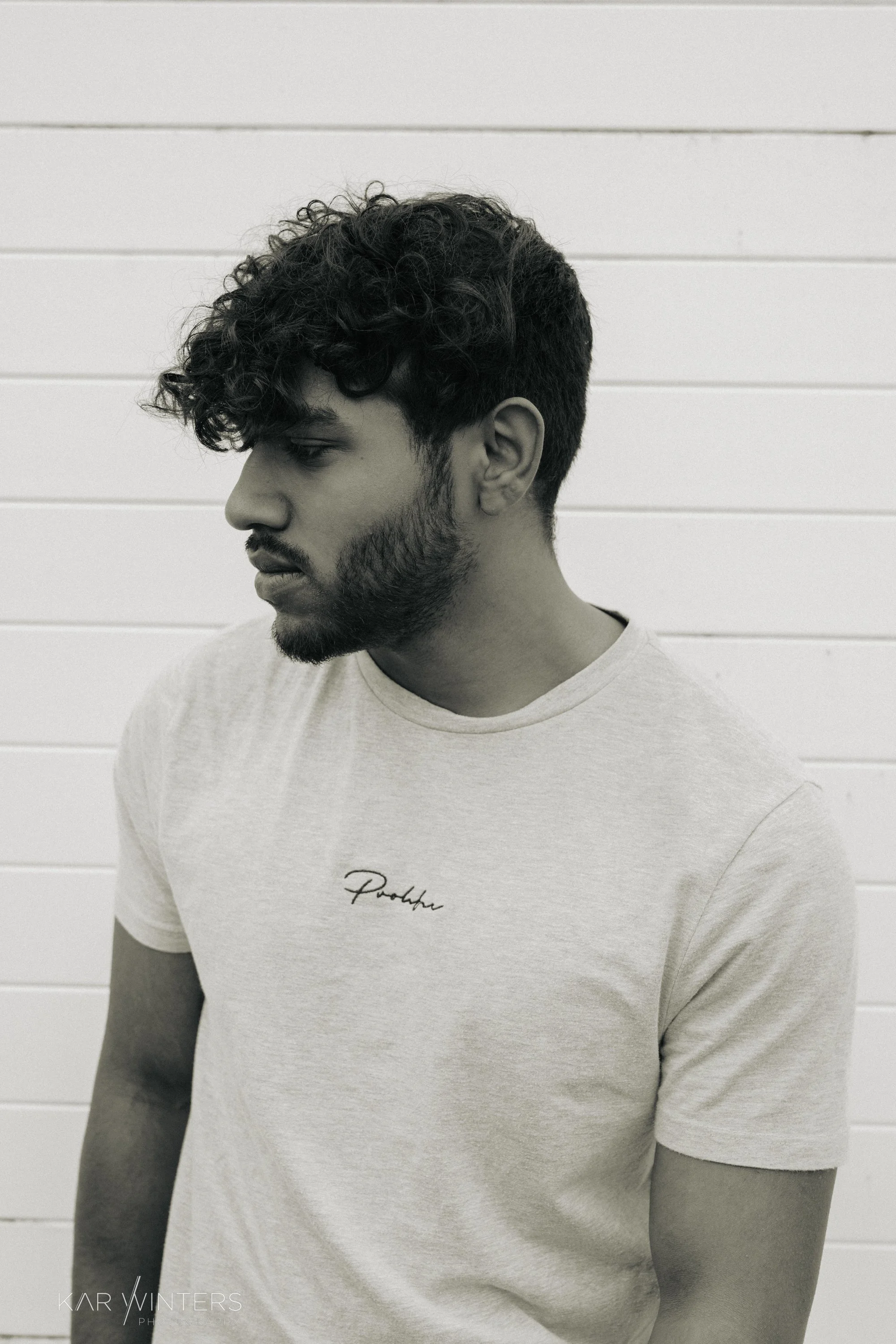 A young man with curly hair and a beard looking down, standing against a white horizontal-paneled wall, wearing a light-colored t-shirt with the word 'Pothe' written on it.