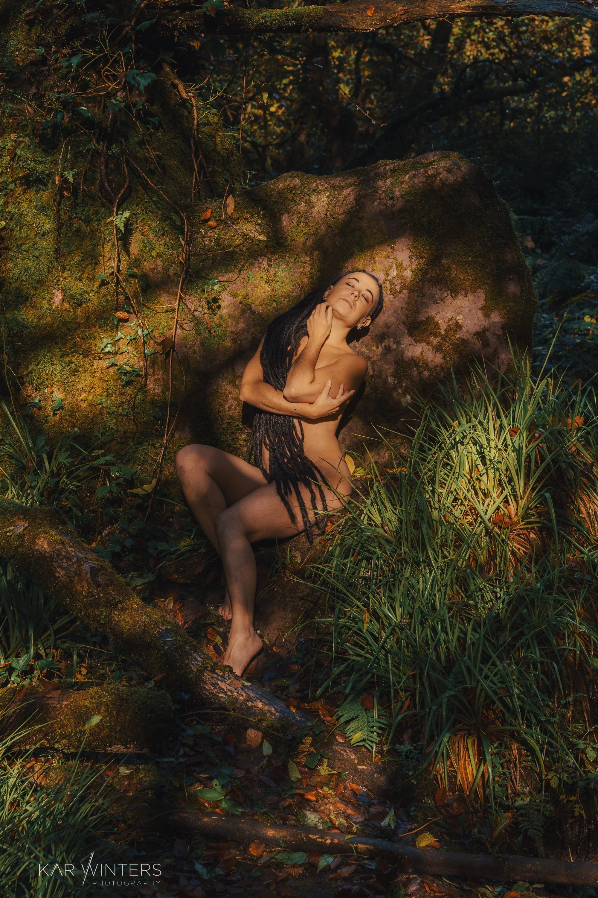 A woman with long dreadlocks posing nude in a forest, sitting on a mossy log, surrounded by greenery and large rocks.