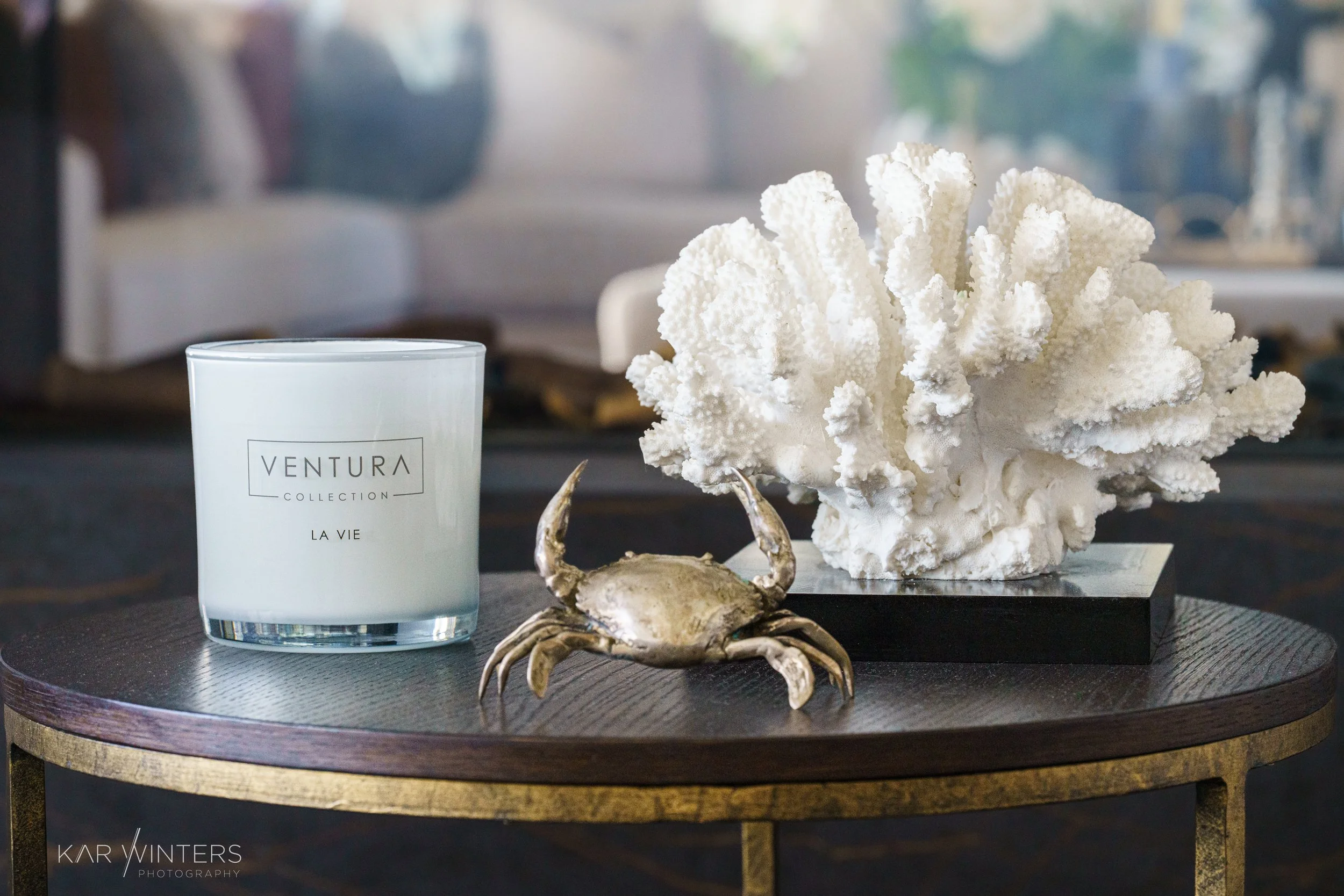 Decorative items on a dark wooden table, including a white scented candle labeled 'VENTURA COLLECTION LA VIE', a metallic crab sculpture, and a large white coral piece on a black stand.