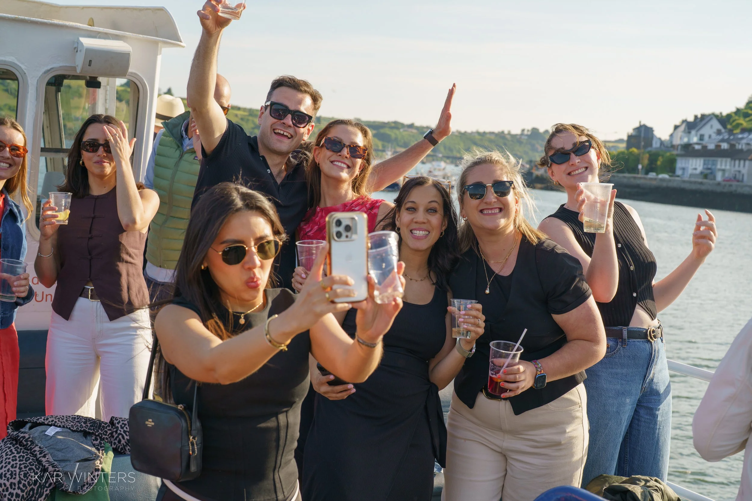 Group of friends on boat taking a selfie, holding drinks, smiling, with water and shoreline in background on a sunny day.