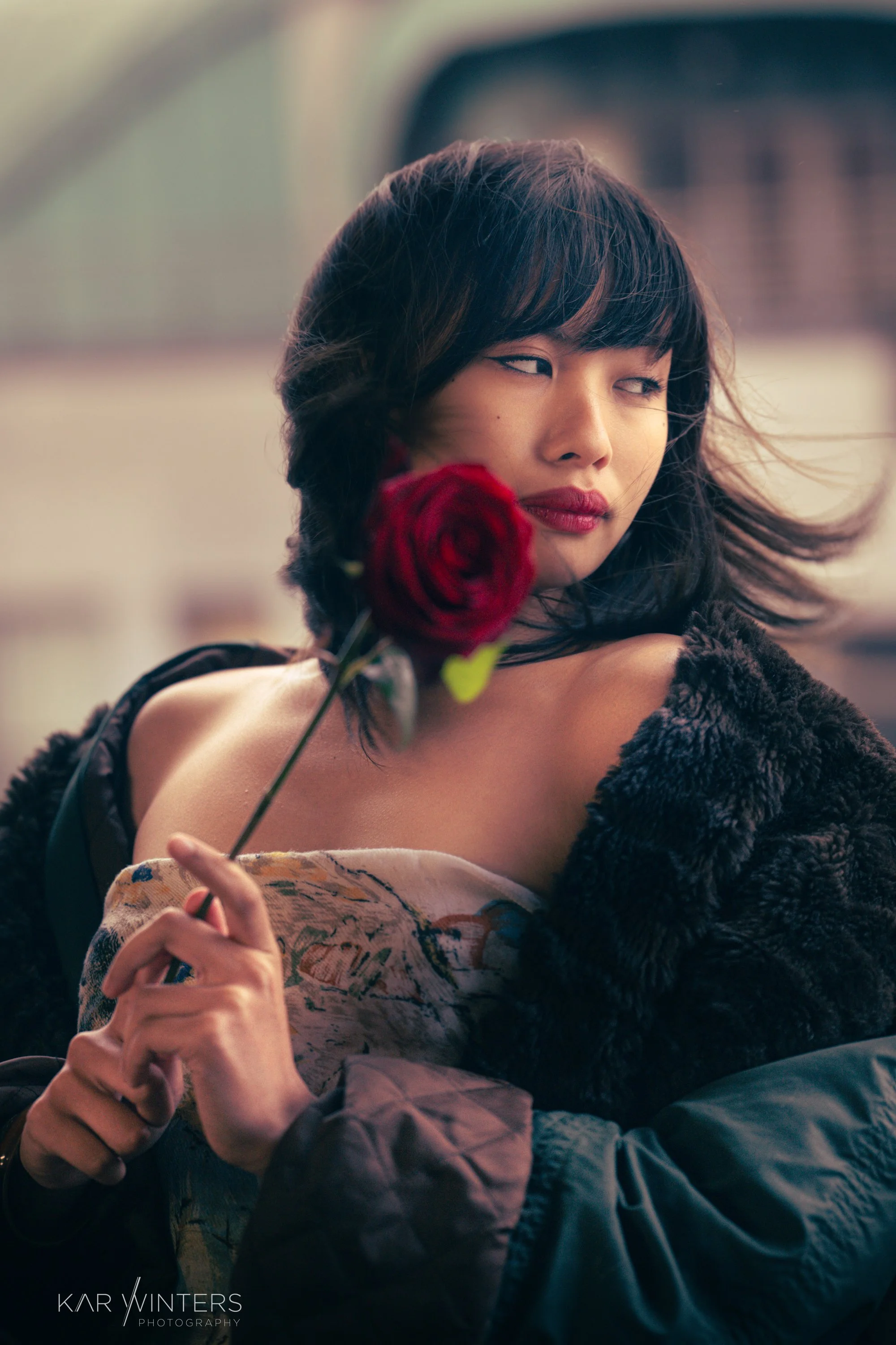 A woman with black hair and red lipstick holds a red rose near her face, wearing a patterned top and a dark faux fur coat, with her head turned slightly to the side, looking away.