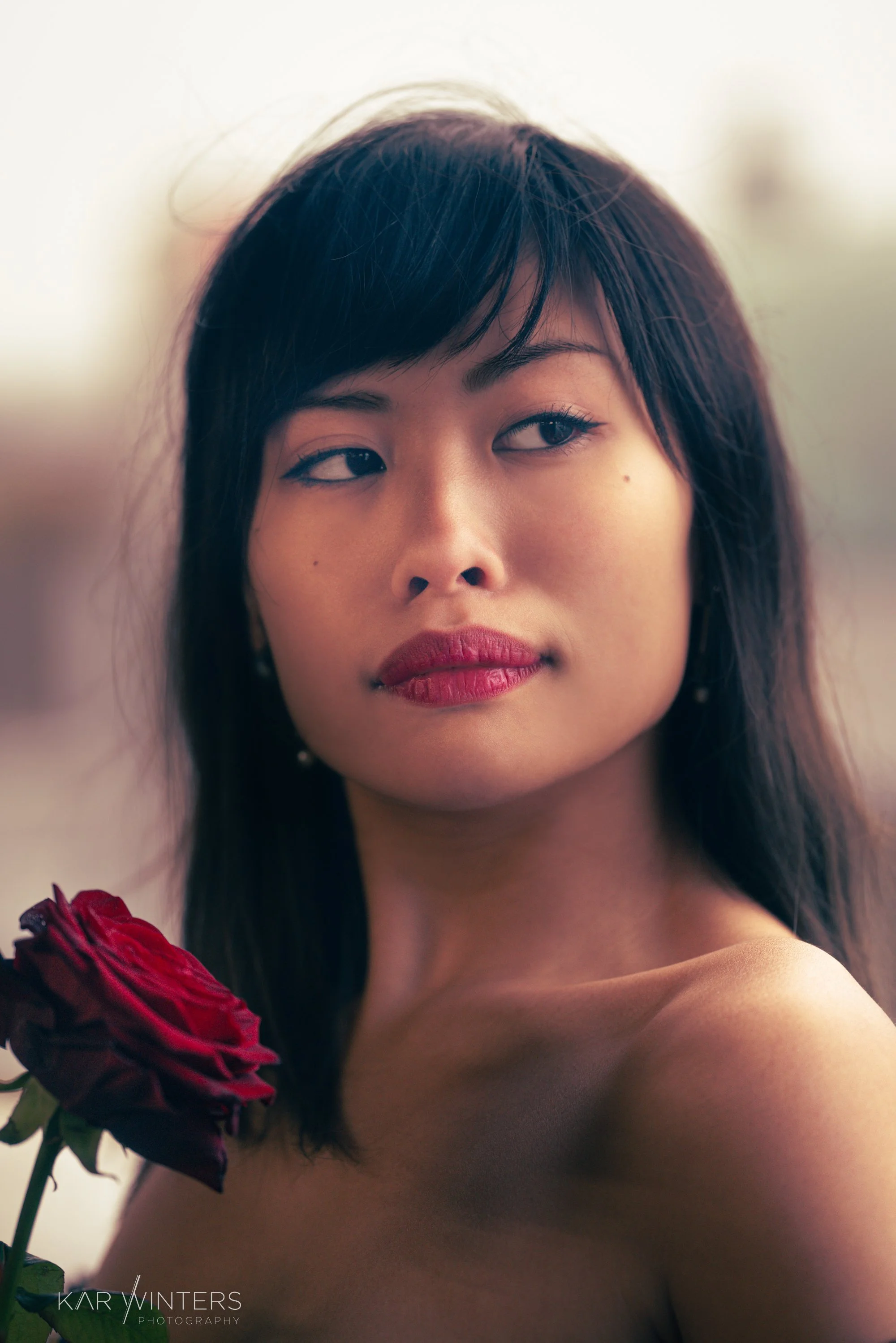 A woman with dark hair and red lipstick holding a red rose near her shoulder, looking to her left with a neutral expression.