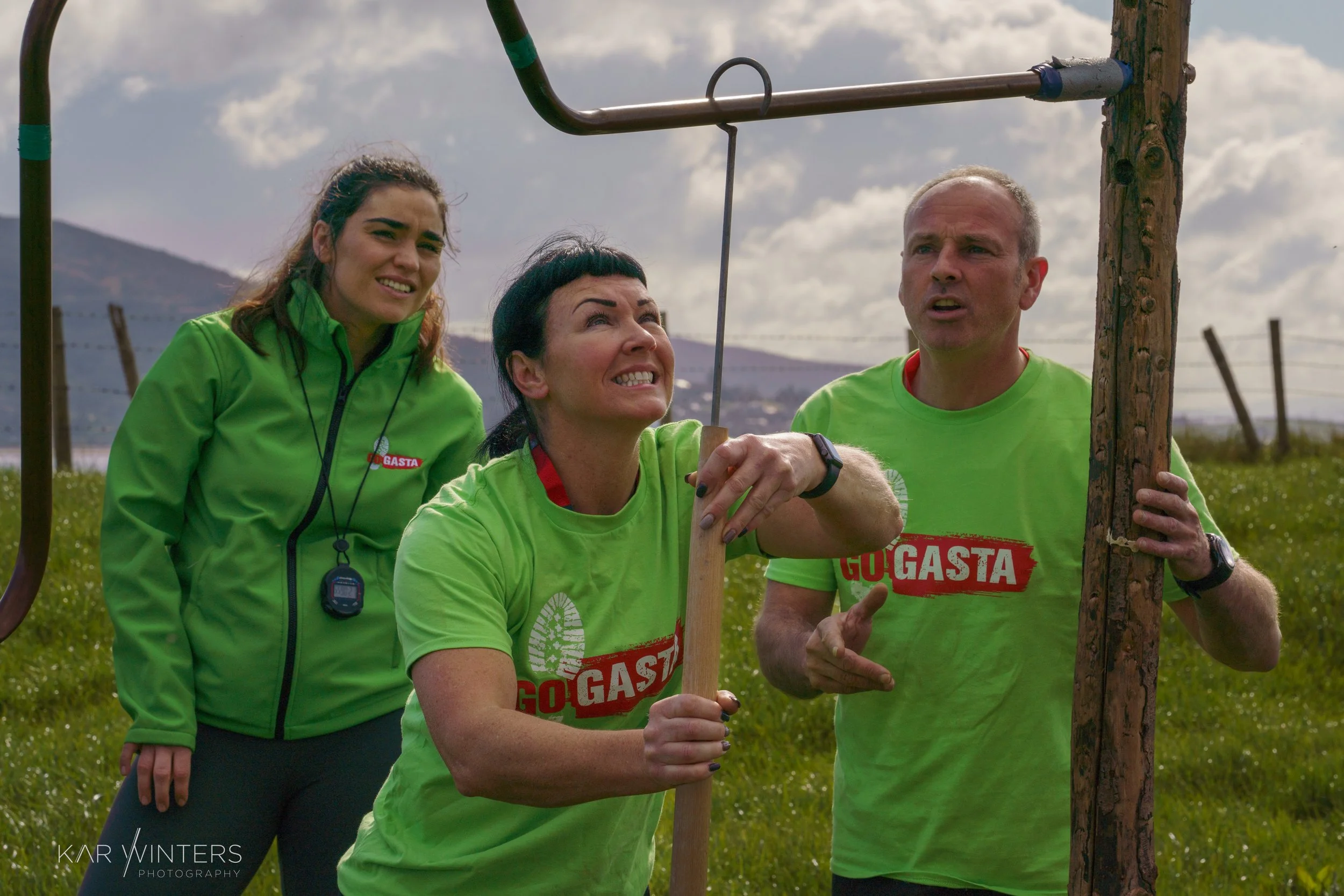 Three people engaging in an outdoor team-building activity. Two are wearing matching bright green shirts with the words 'GO GASTA' and one woman is wearing a green jacket with a whistle. They are holding equipment, and one woman is trying to climb or
