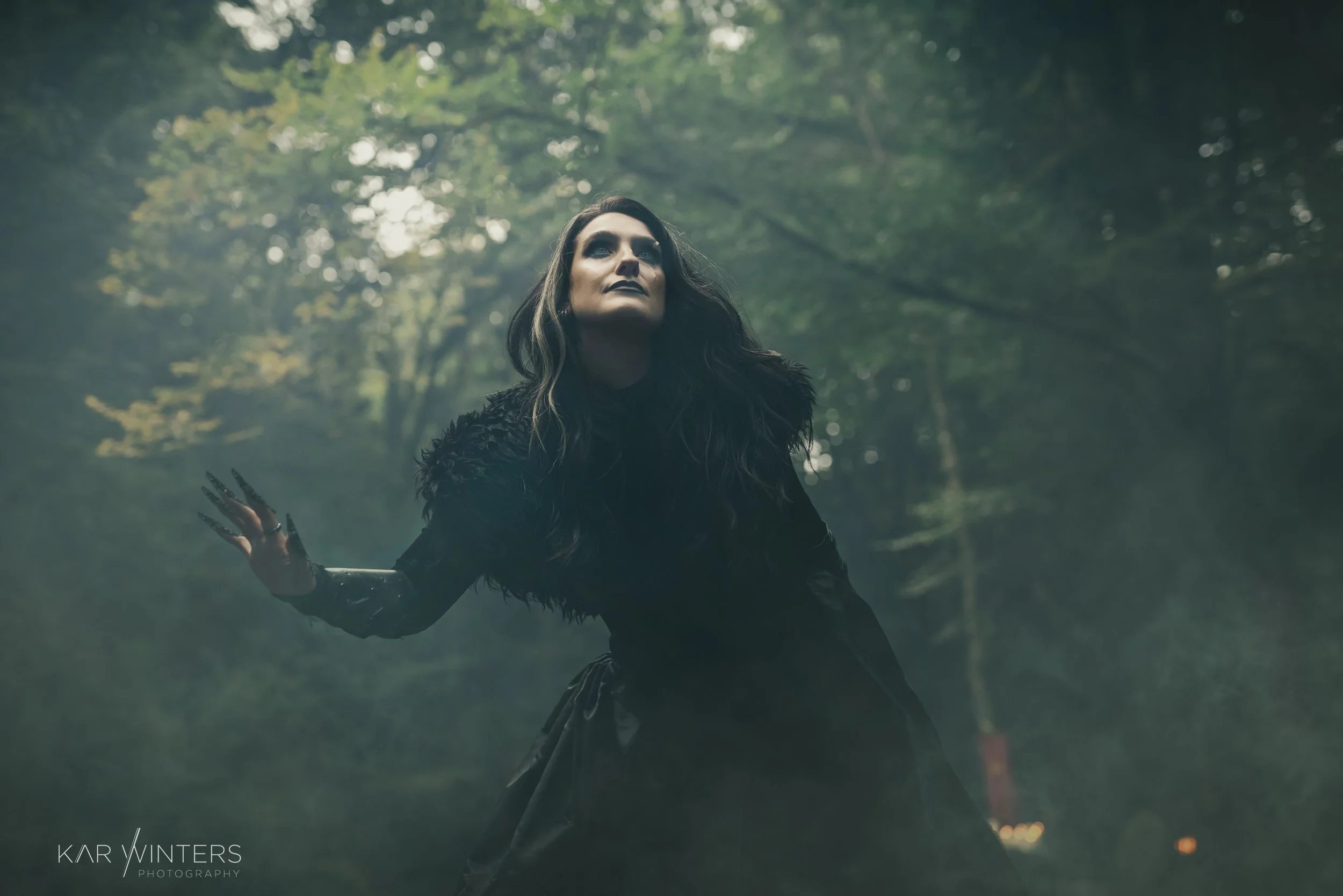 A woman dressed in black with long dark hair outdoors in a forest, with trees and sunlight filtering through the leaves.