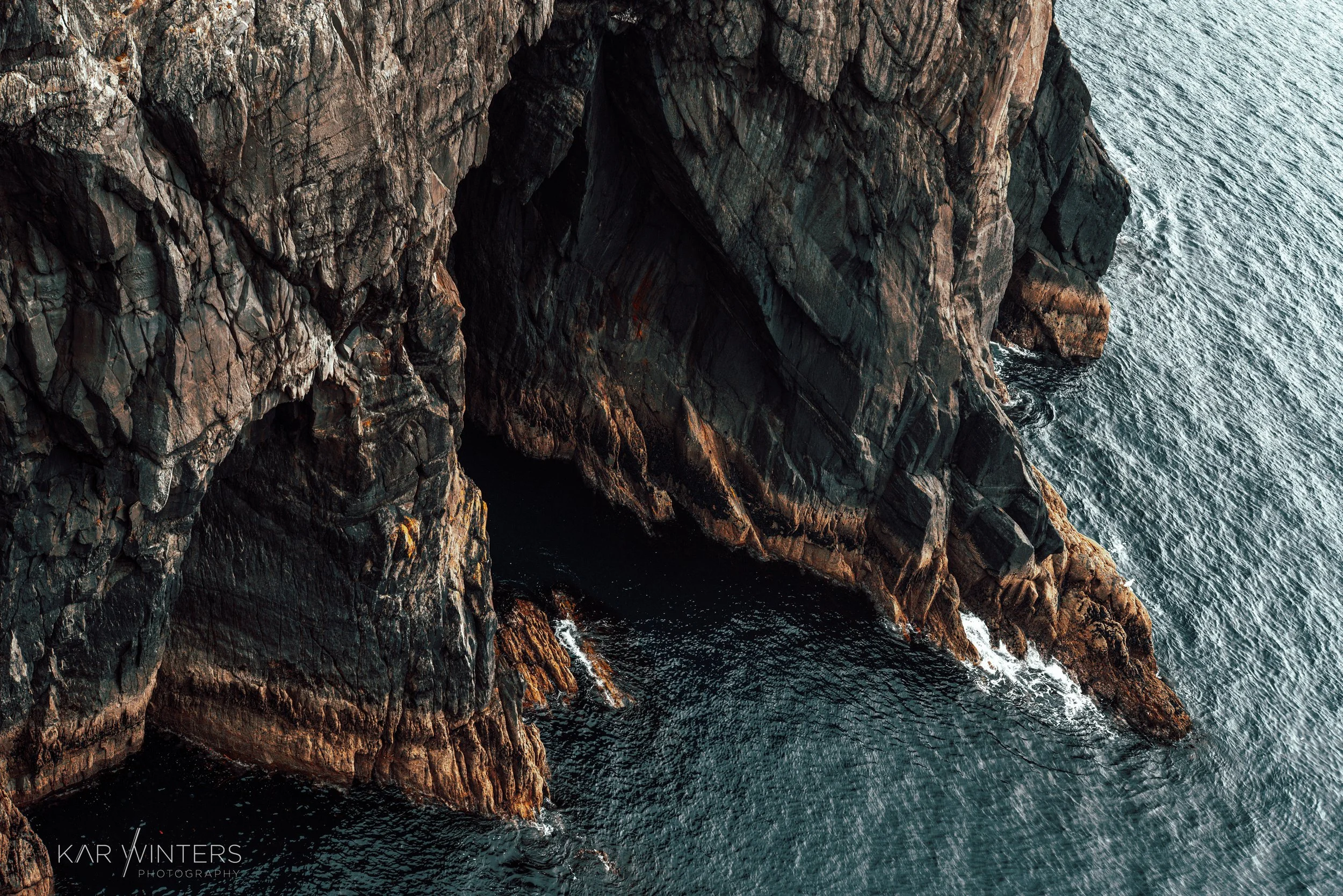 Aerial view of rugged rocky cliffs rising from the ocean, with waves crashing against the base of the rocks.