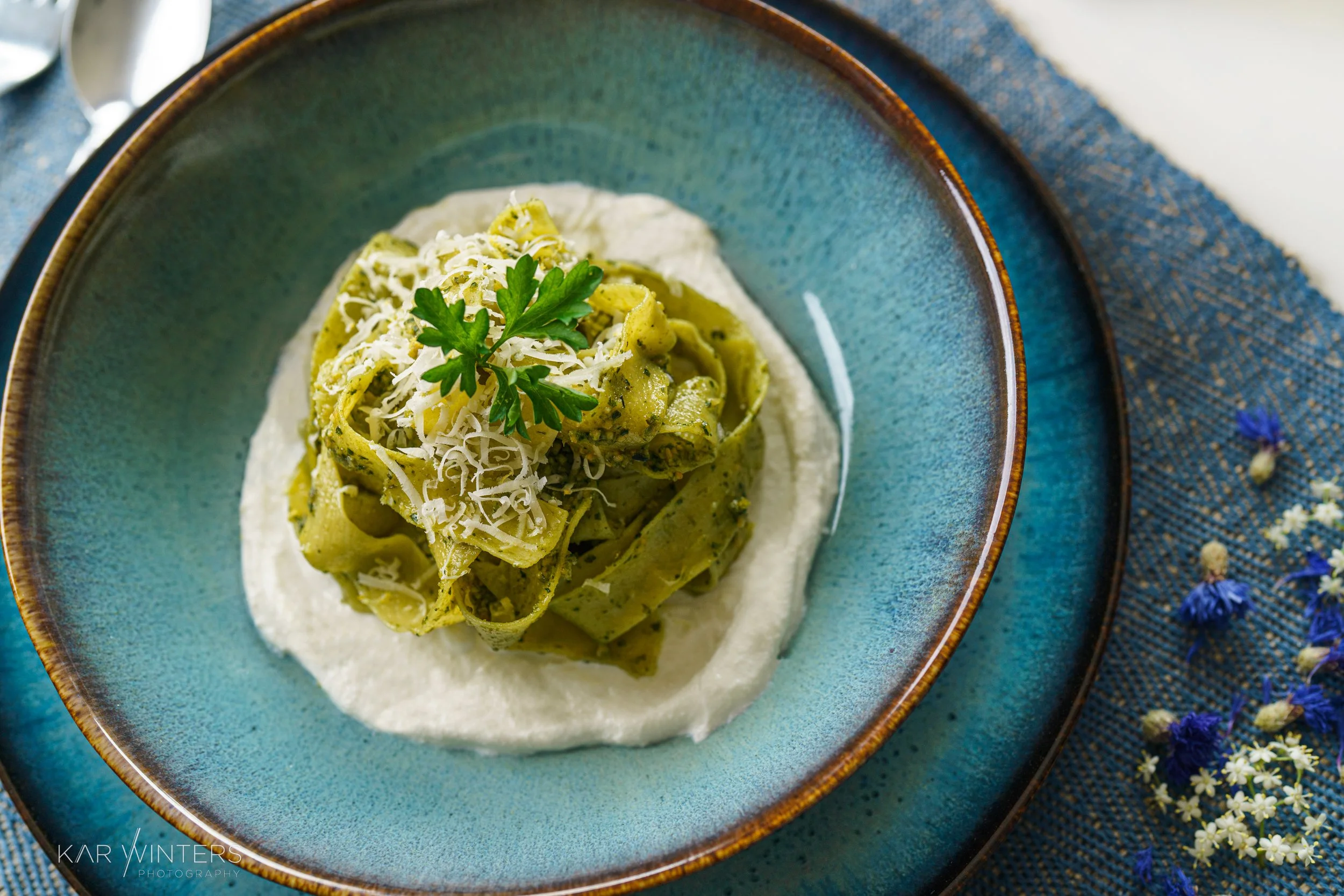 Plate of green spinach and basil pesto fettuccine topped with grated cheese and a sprig of parsley, served on a blue ceramic plate with a cream sauce on the side, on a blue cloth with small flowers.