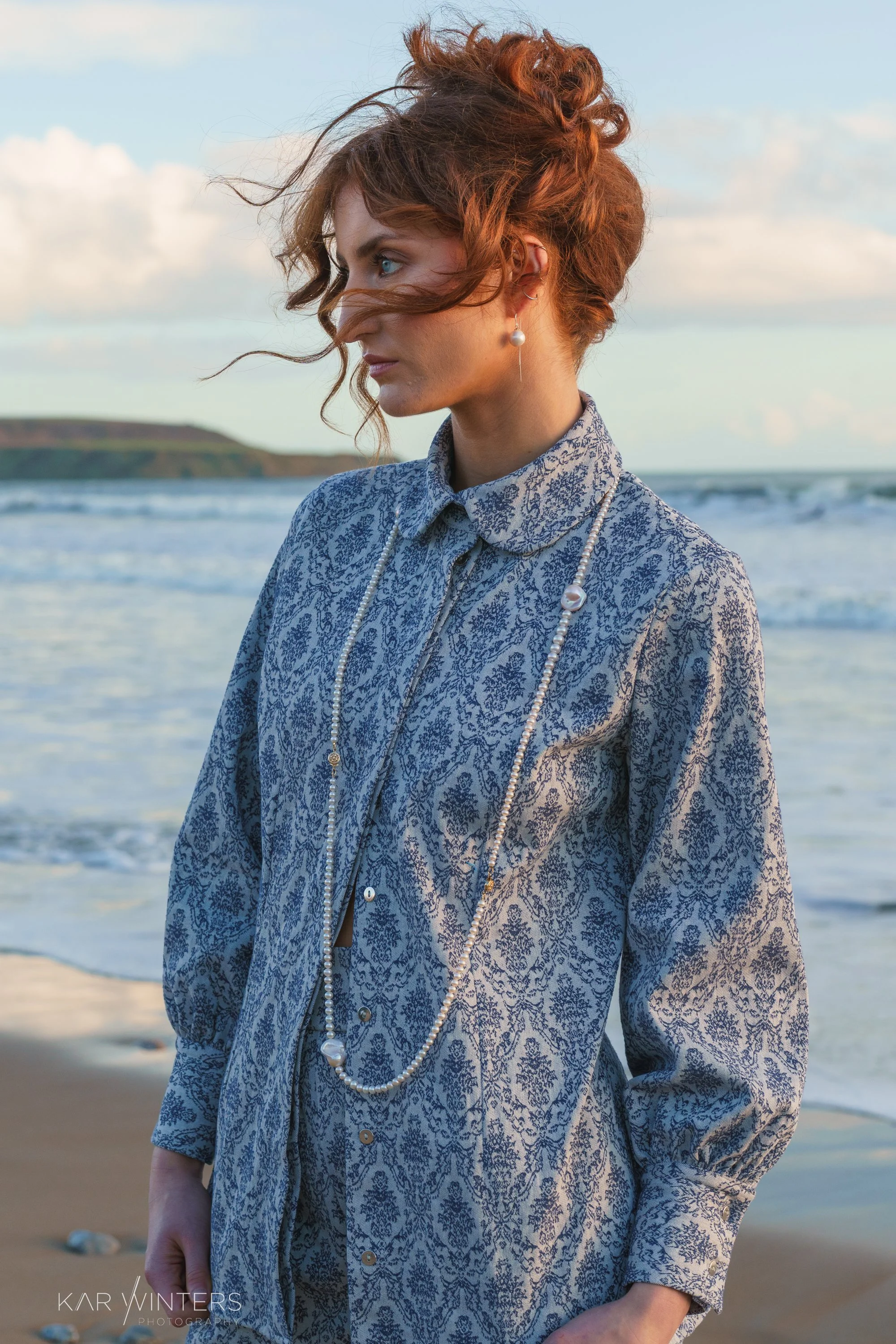 A woman with red hair and blue eyes stands on the beach in a long-sleeved, patterned shirt wearing pearl earrings and necklaces, with the ocean waves and a distant green hill in the background.