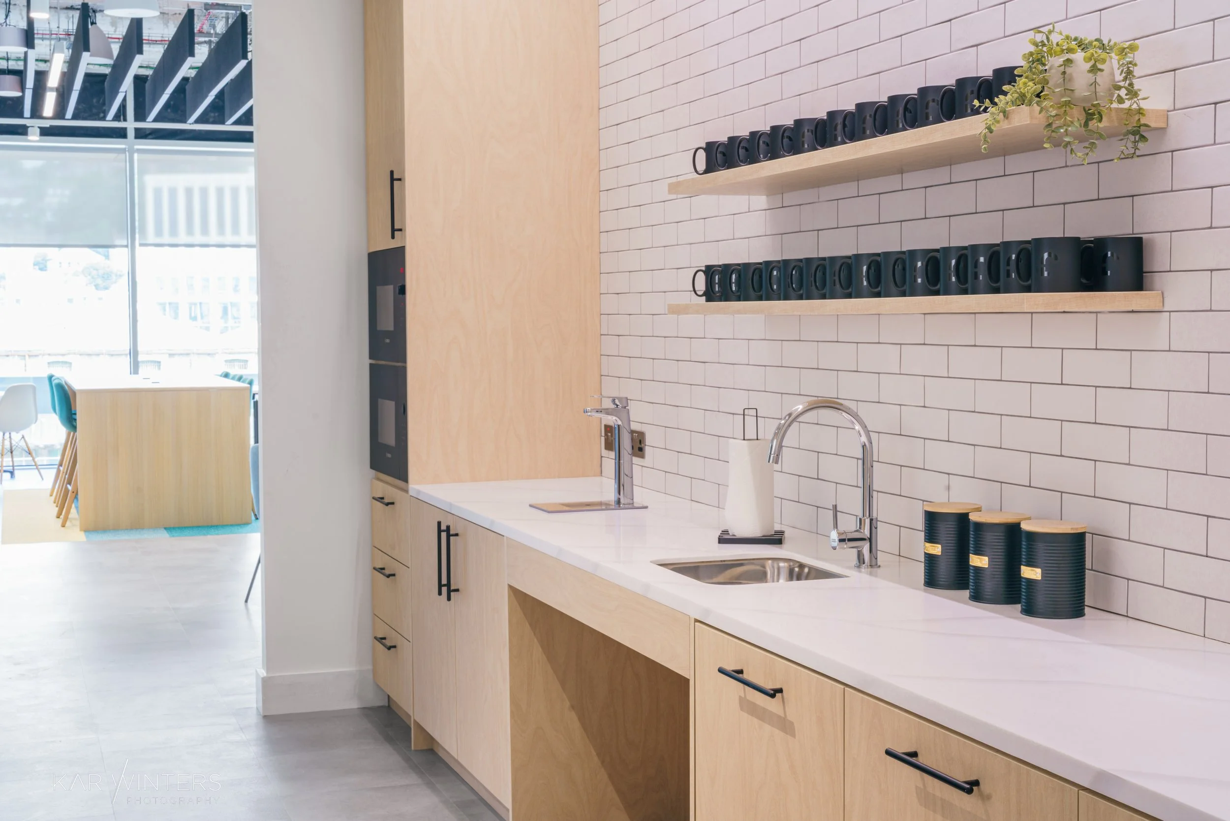 Modern kitchenette with white subway tile wall, wooden cabinets, black mugs on floating shelves, and a stainless steel sink and faucet.