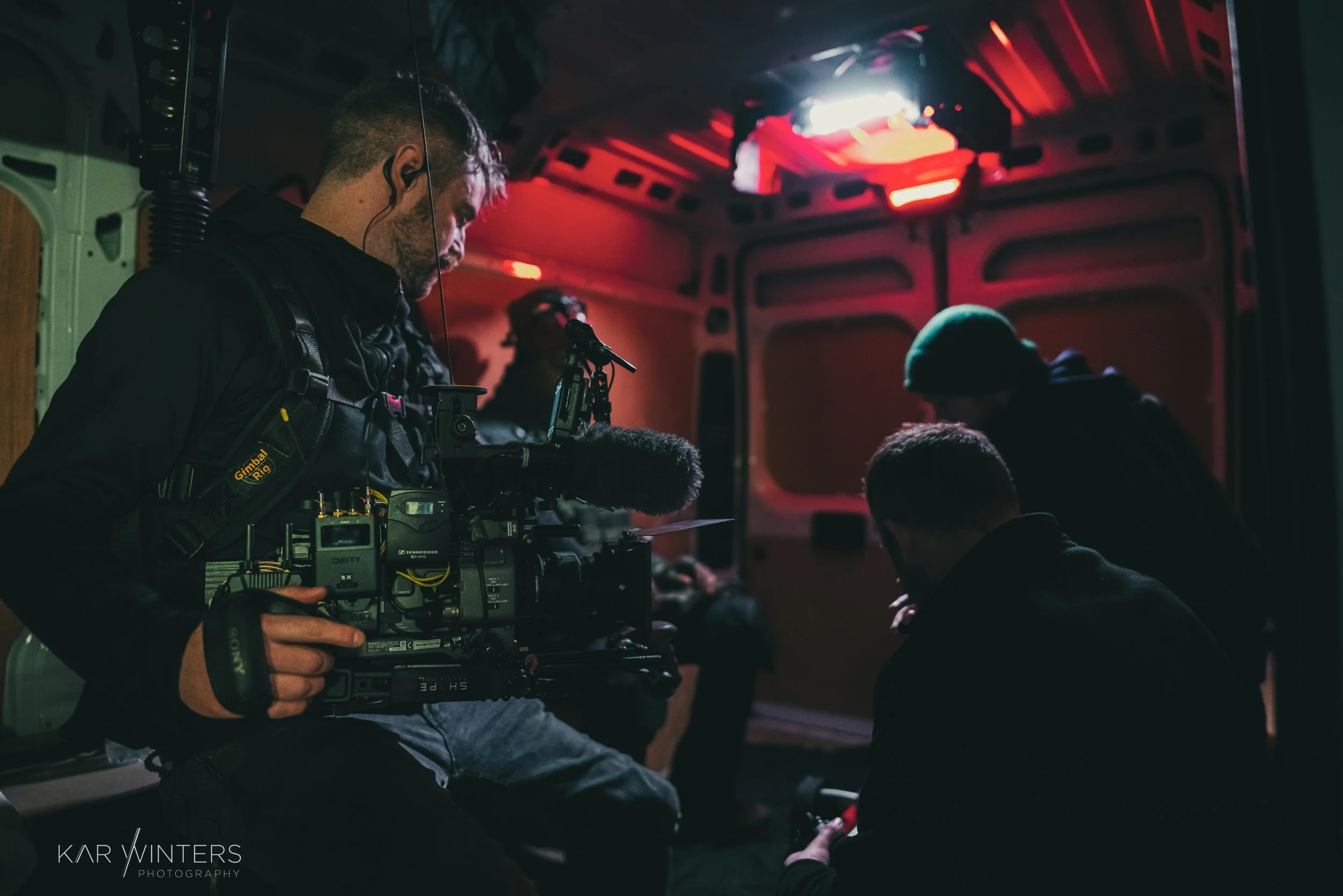 Film crew inside a vehicle, with one person operating a professional camera, others gathered around, illuminated by dim red and white lighting.