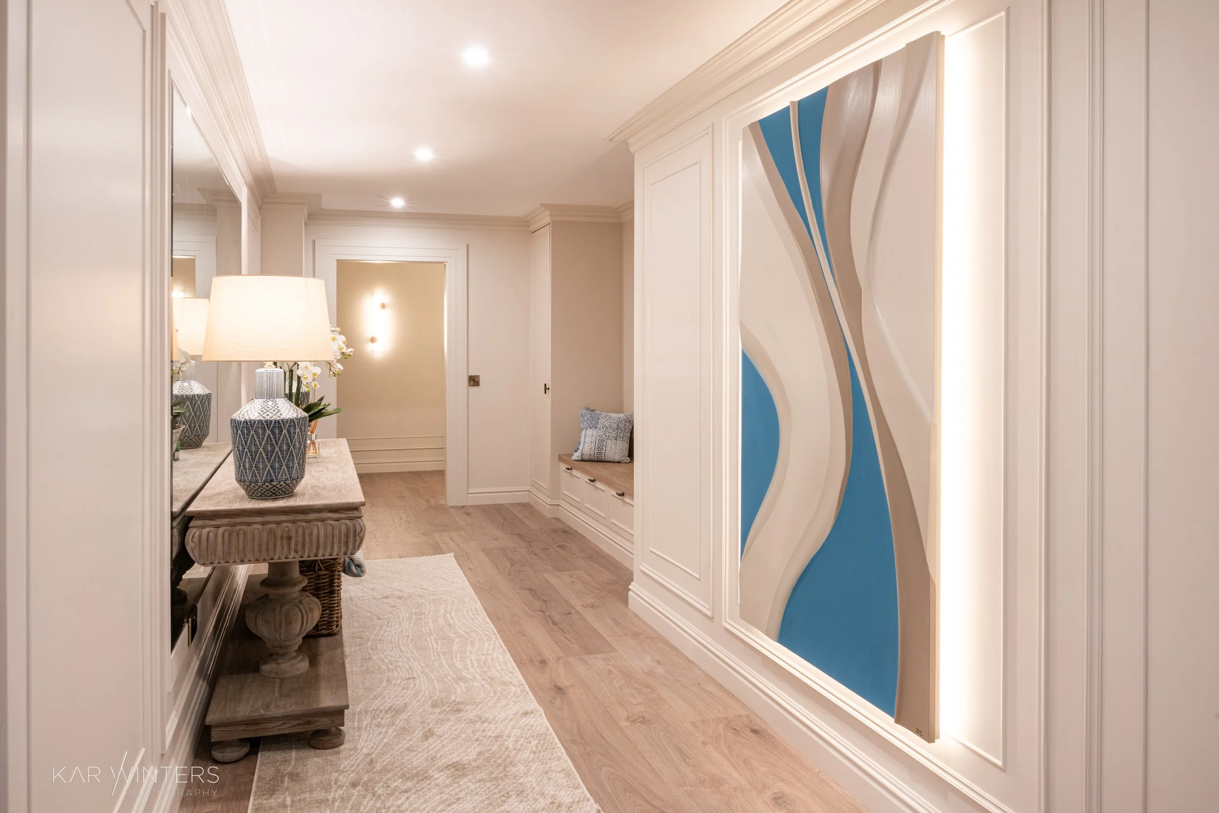 Elegant hallway with white walls, hardwood flooring, and recessed ceiling lights. Features a console table with decorative lamps and vases, a built-in bench with pillows, and a modern colorful abstract wall art piece.