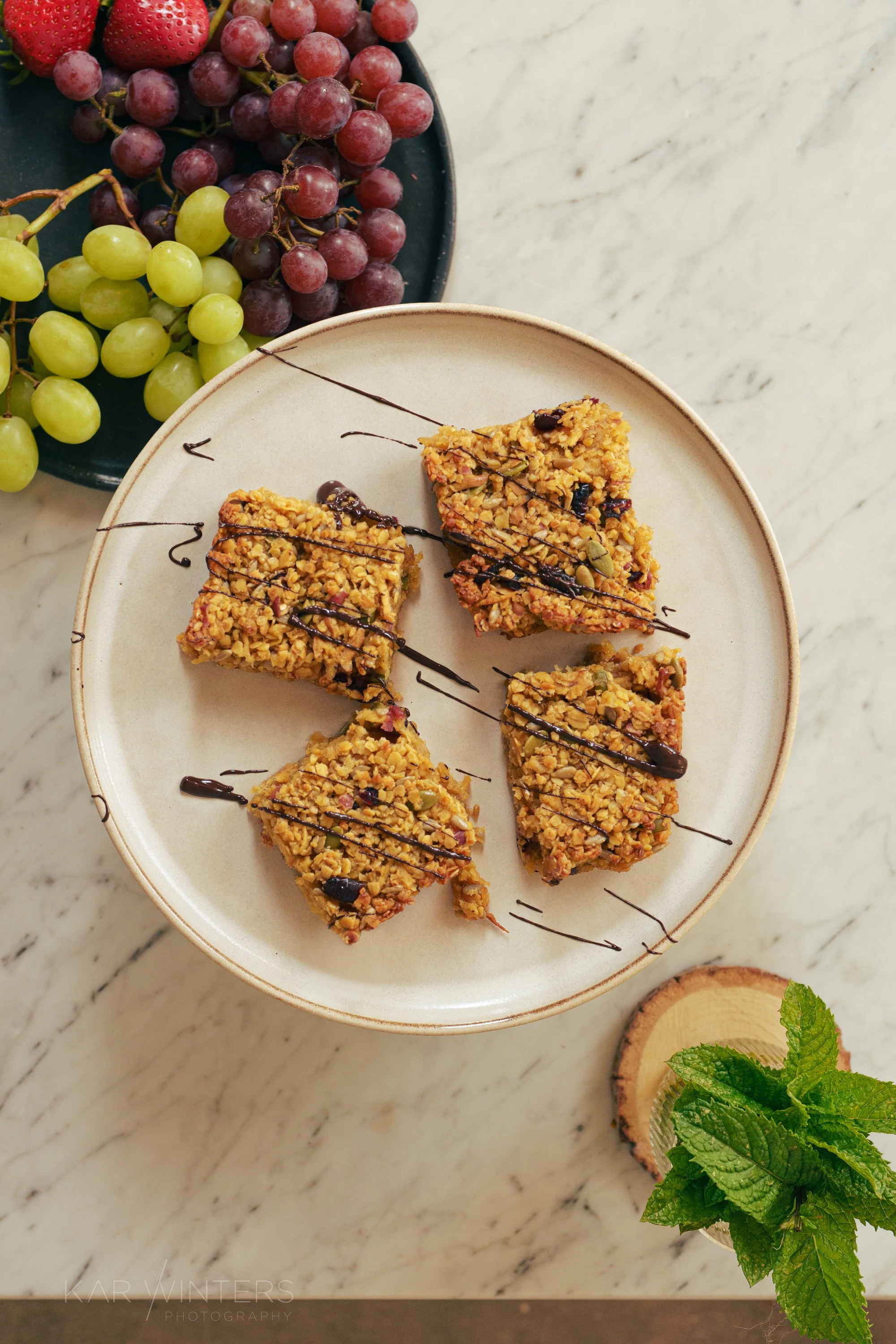 Granola bars with drizzle on a white plate, grapes and strawberries on a black plate, and fresh mint leaves in a glass on a marble surface.