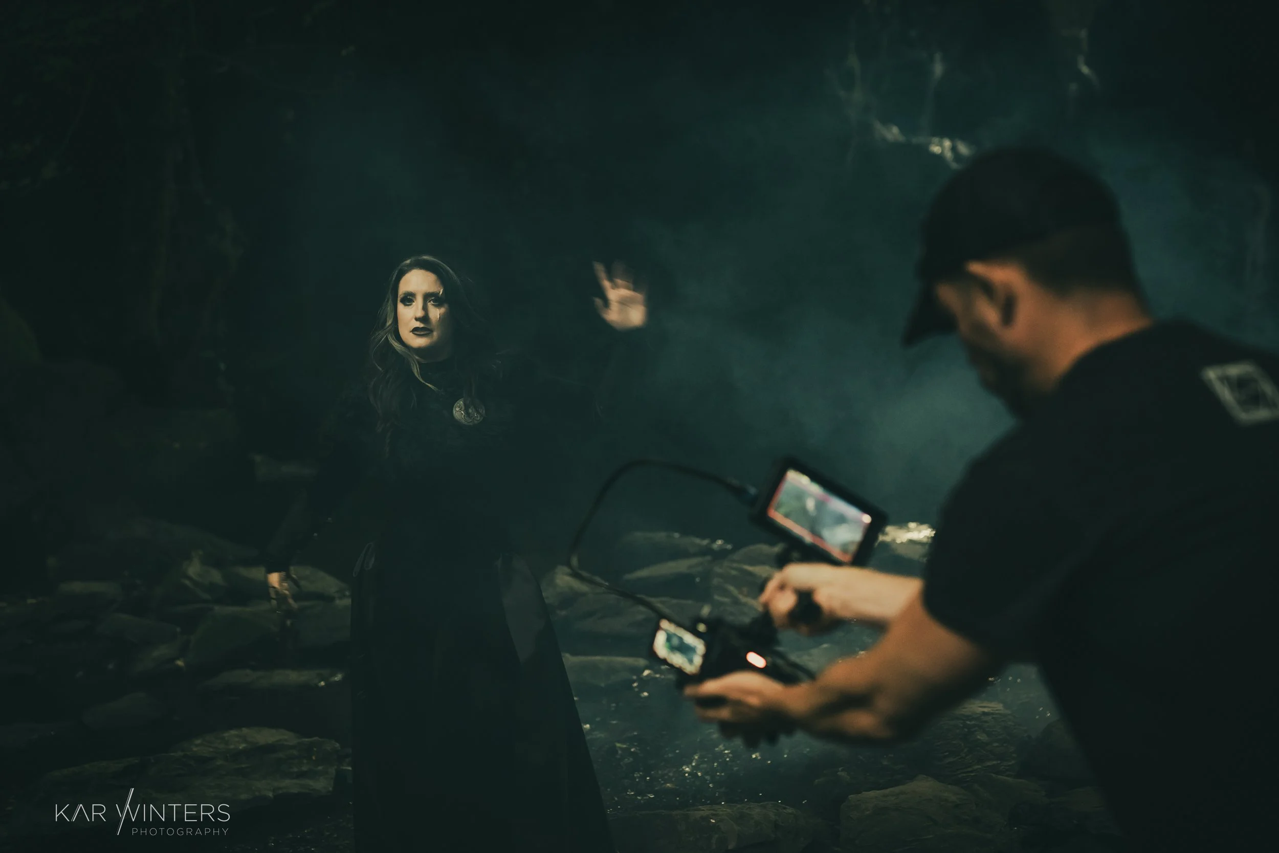 A woman in goth attire standing in a dark, misty forest while a man takes her photograph with a camera.
