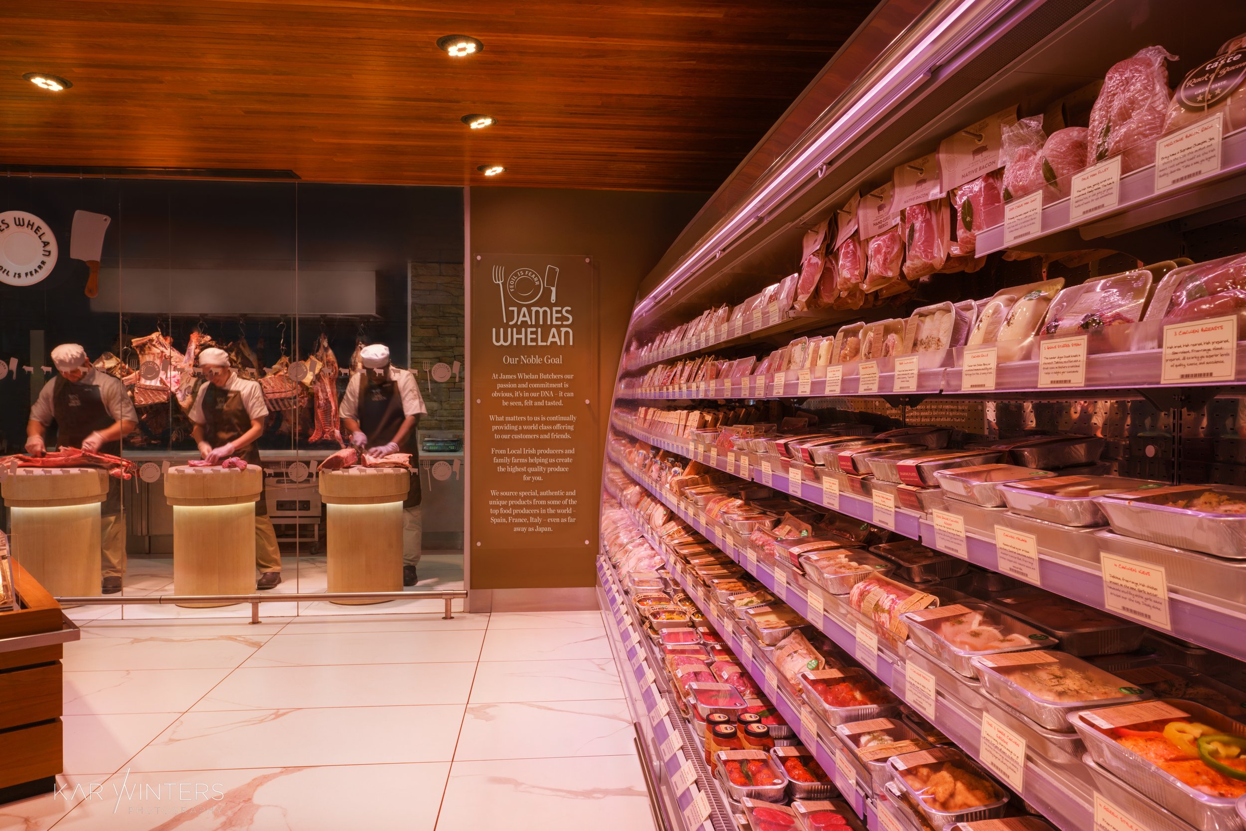 Employees in chef hats and aprons preparing meat behind a glass window at James Whelan Butchers, with shelves of packaged meats on the right.
