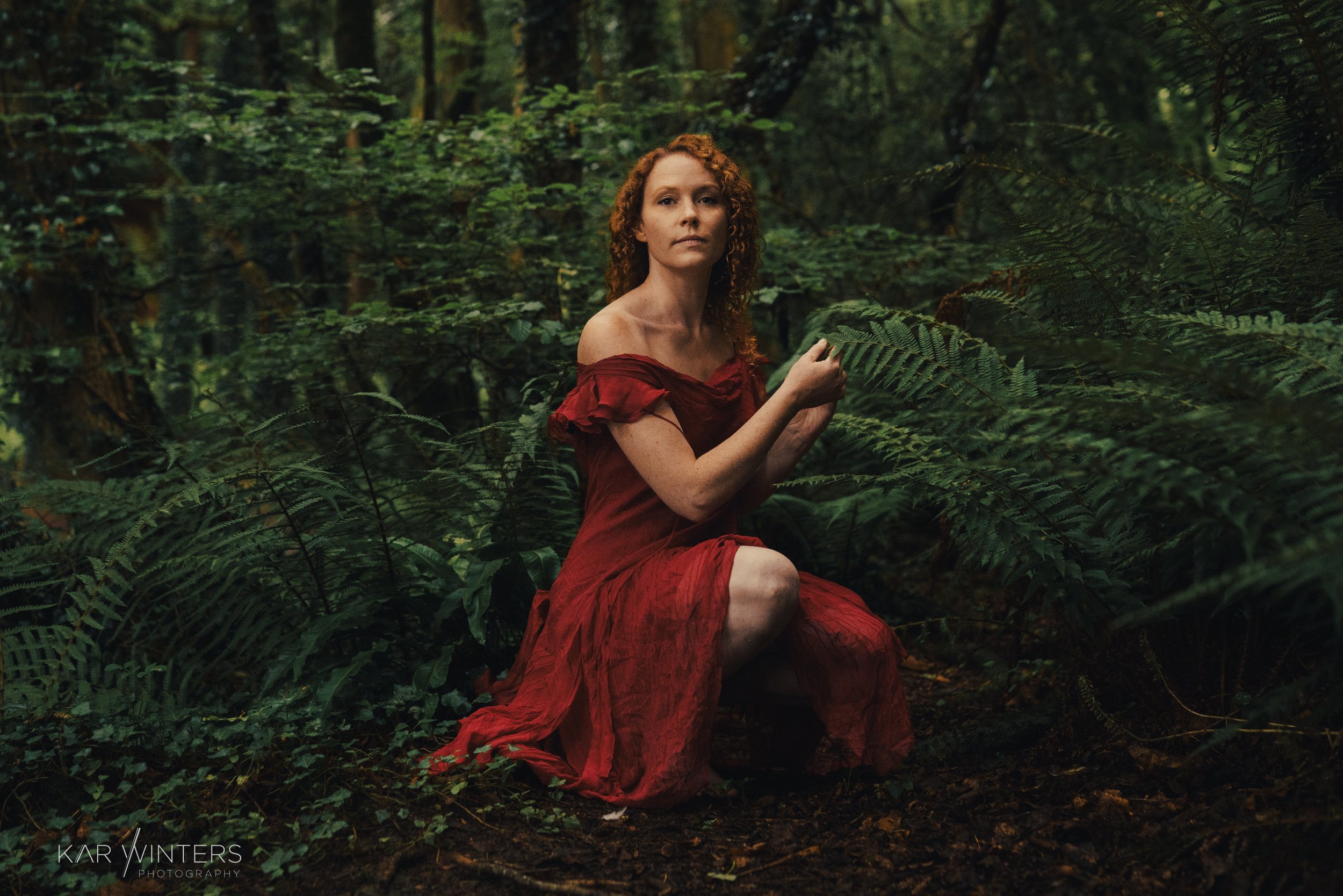 A woman with red curly hair, wearing a red off-shoulder dress, kneels in a lush, green forest surrounded by ferns.