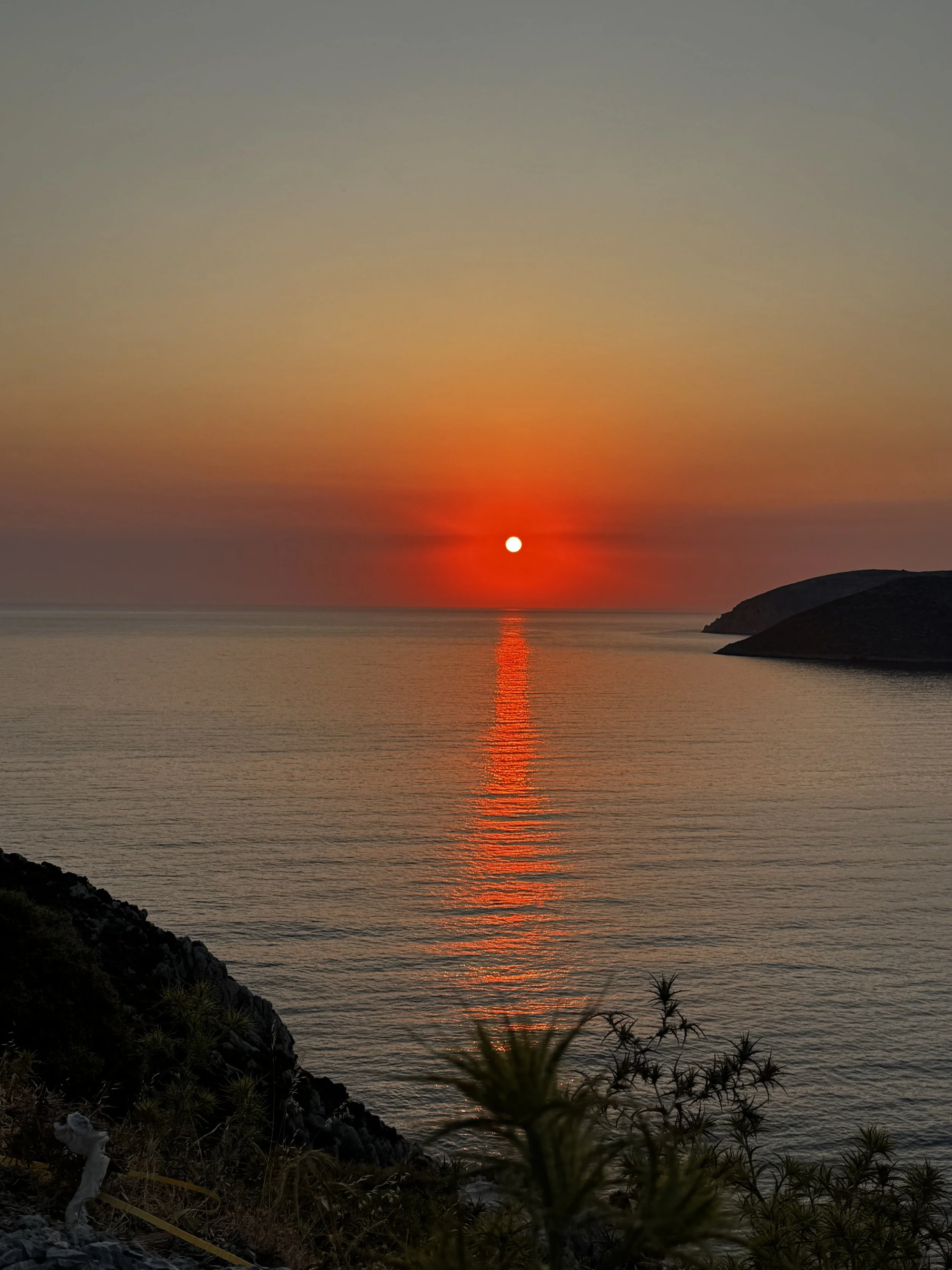 A sunset over the ocean with the sun near the horizon, casting orange and red colors in the sky and reflecting on the water. A dark hill is visible on the right, and some plants and rocks are in the foreground.
