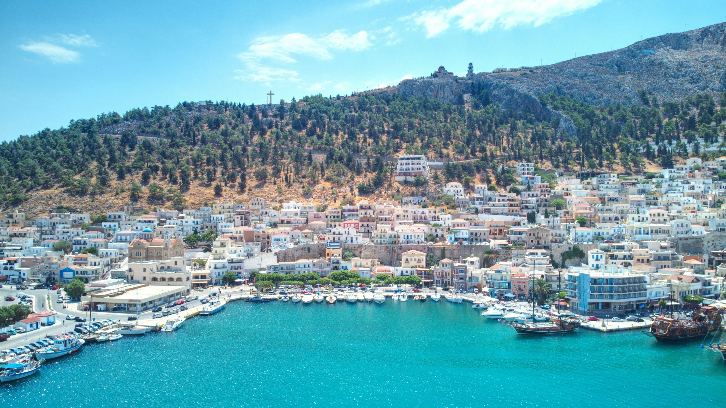 Scenic view of a coastal city with white buildings, a marina, and boats under a cloudy sky, hillside with trees, and a cross on a hill in the background.