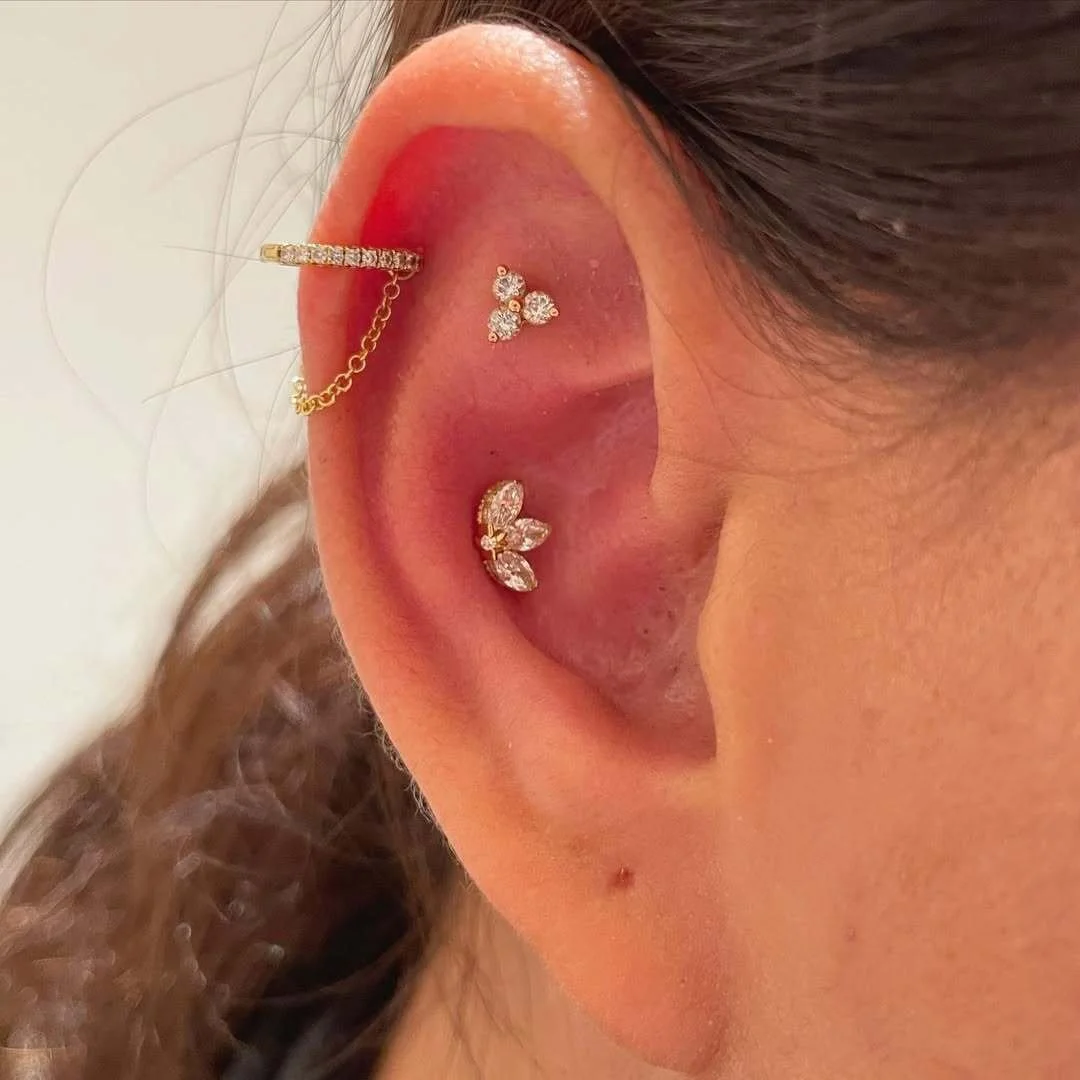 Close-up of an ear with multiple piercings, featuring gold and jeweled earrings. A small hoop with a chain, a crystal stud, and a cluster of marquise-shaped stones are visible.