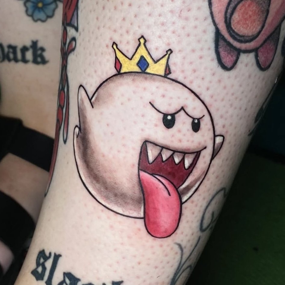 Tattoo of a cartoon ghost character with a crown, sticking out its tongue, on a person's skin.
