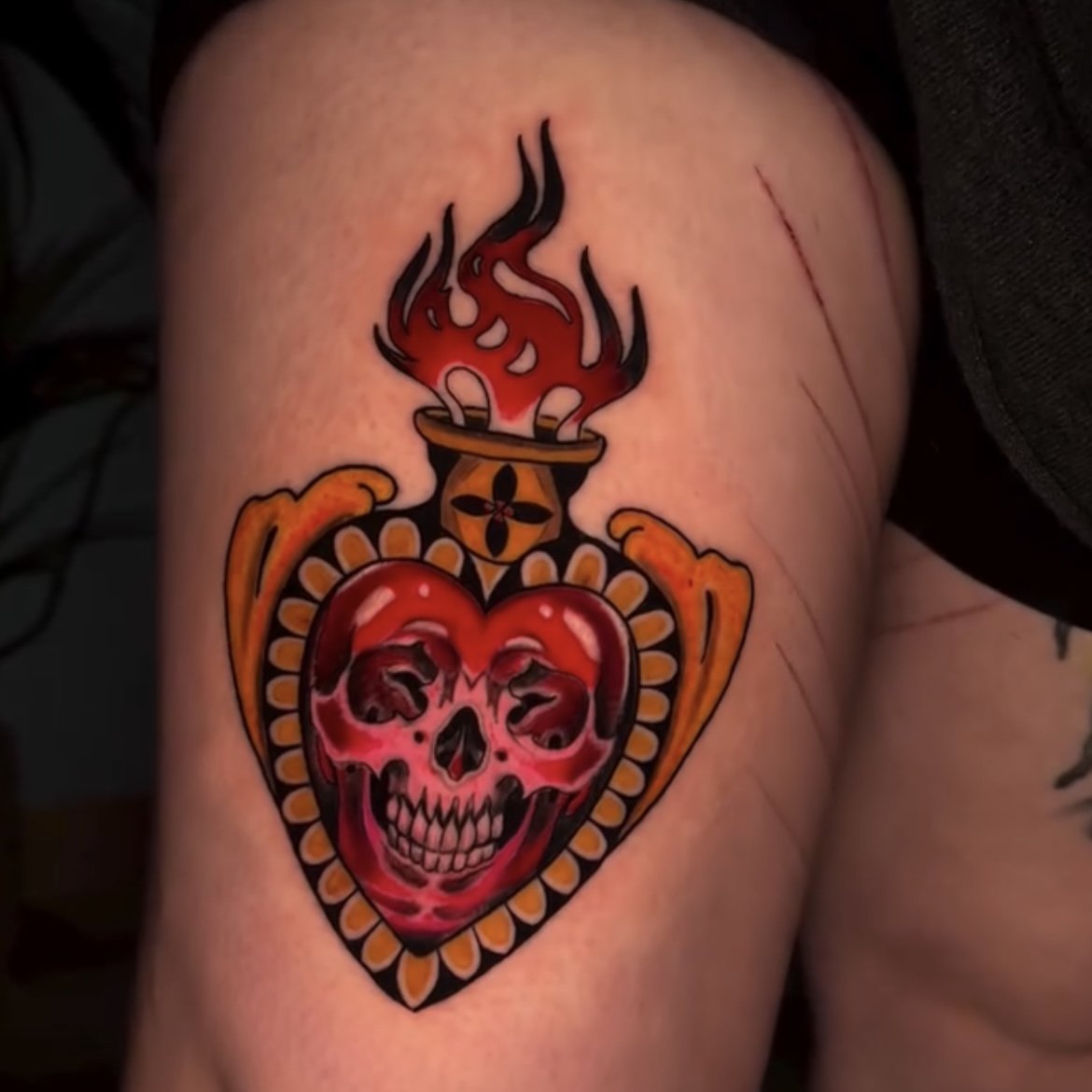 Tattoo of a flaming heart with a skull inside, embellished with yellow details, on a person's skin.