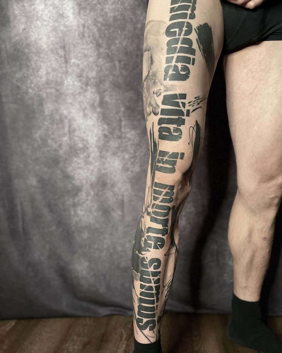 Person's leg with a large tattoo featuring Latin text covering the length of the limb, against a neutral background.